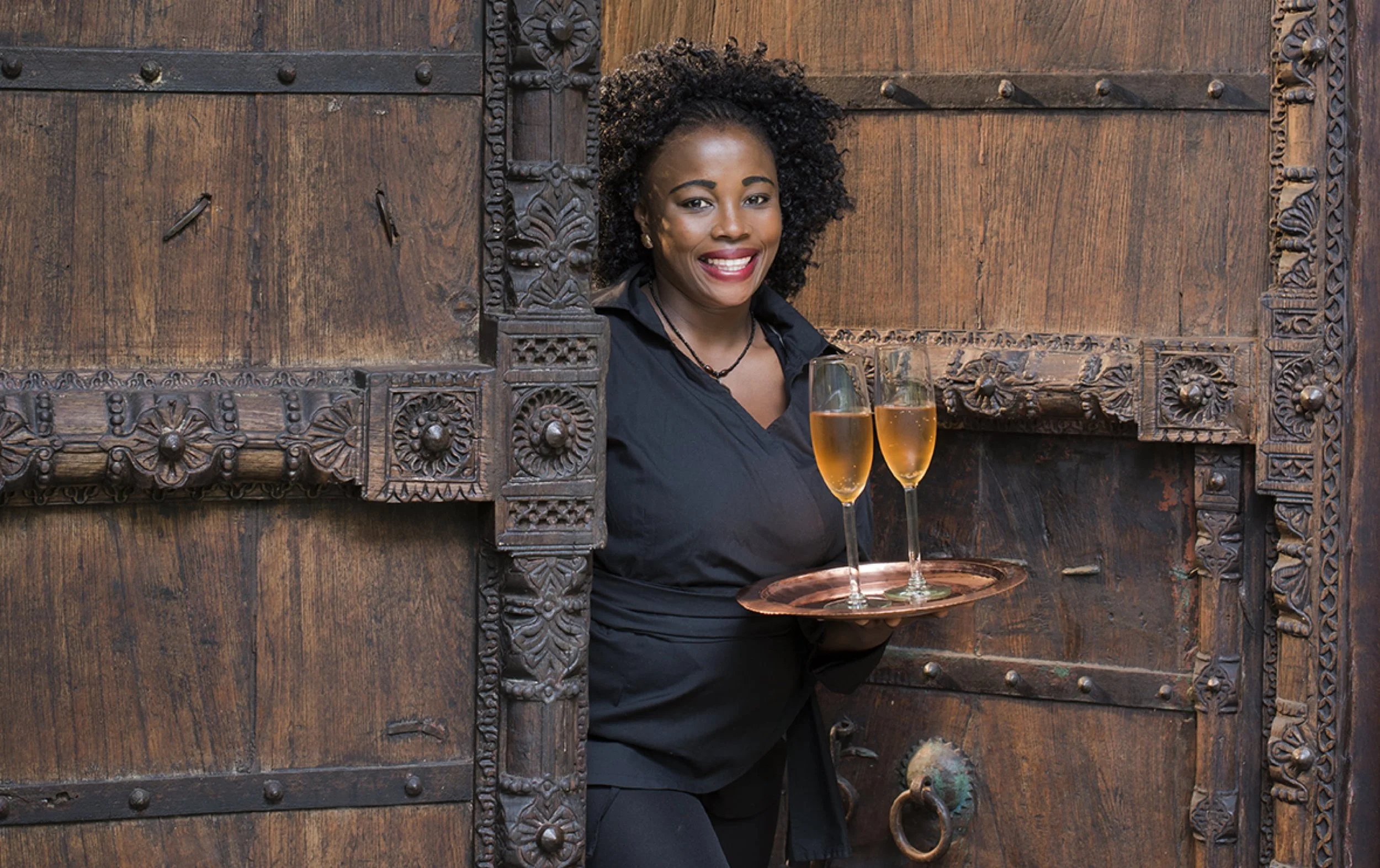 A smiling woman with curly hair holding a tray with two glasses of champagne, standing in front of a large wooden door with intricate carvings.