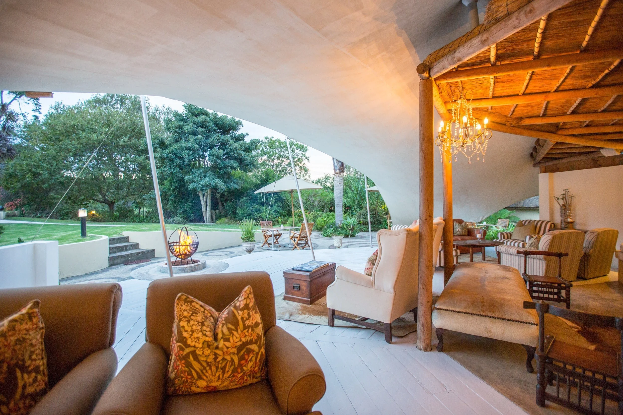 Covered outdoor patio with couch, armchair, wooden furniture, chandelier, and view of garden with trees, potted plants, and a fire pit.