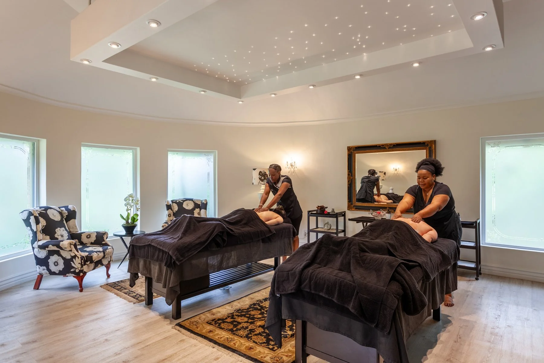 Two massage therapists are giving massages to clients lying face down on massage tables in a well-lit, elegant room with three windows, black-covered massage tables, and floral armchairs.