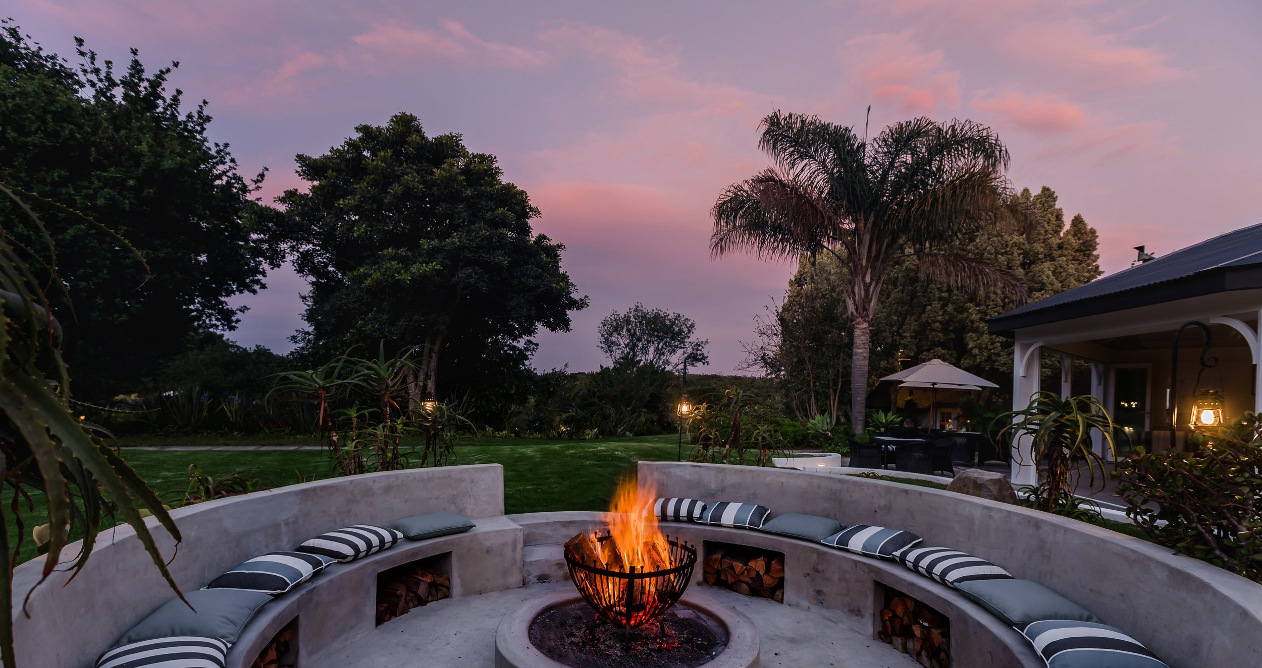 Outdoor backyard with a fire pit surrounded by curved built-in seating with striped cushions, lush trees, a lawn, and landscape lighting, under a pink and purple sunset sky.