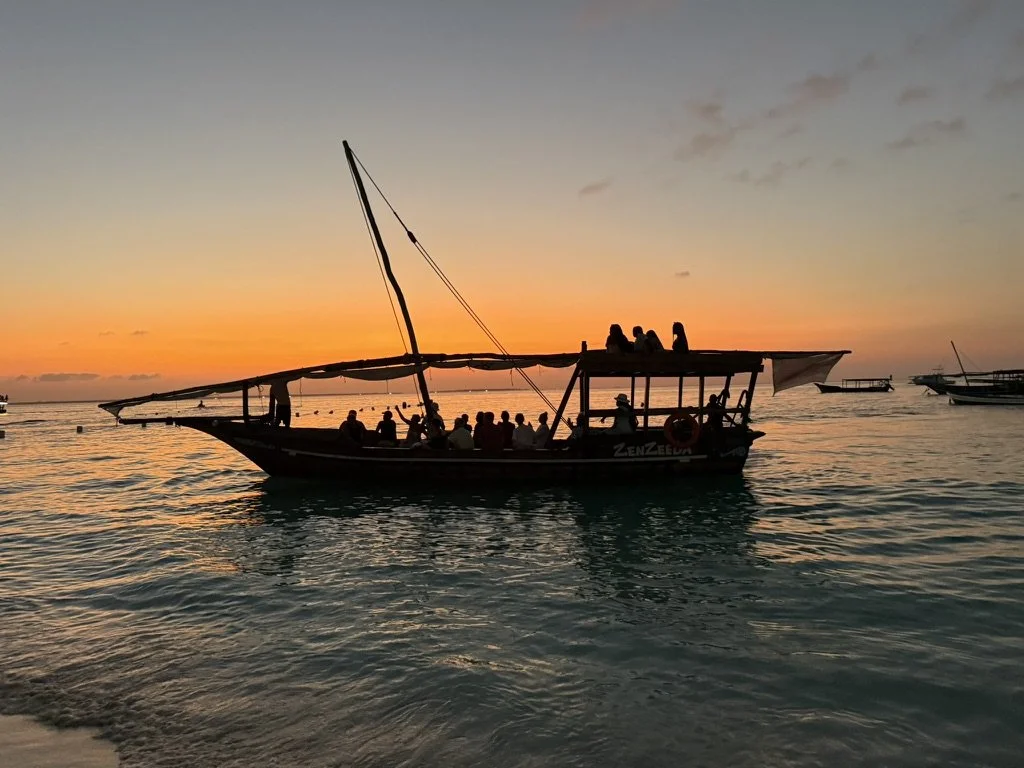 Silhouette of a boat at sunset with people on board, calm ocean water, and a colorful sky.
