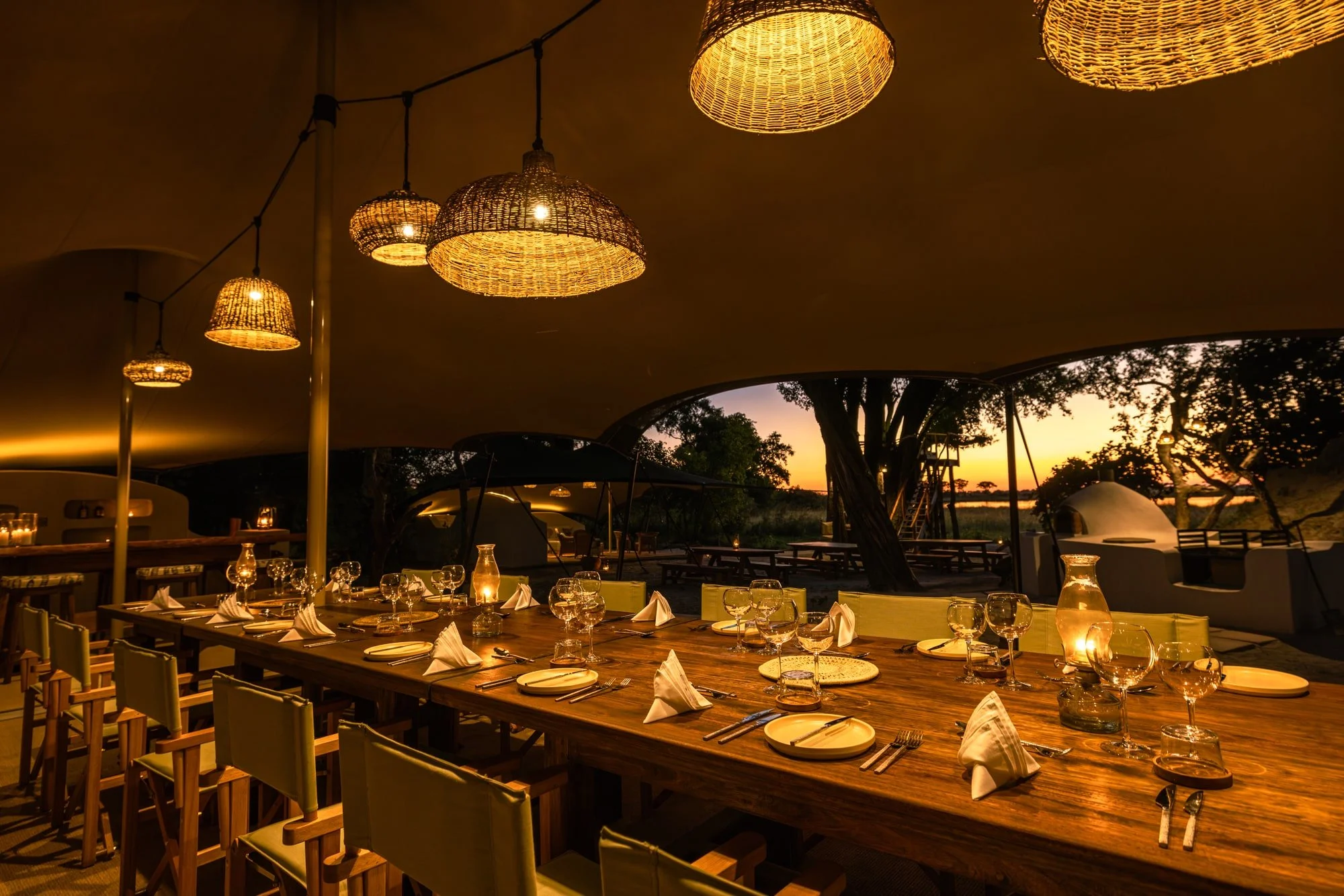 An outdoor dining area under a large canopy with hanging wicker lamps, set for a meal with glasses, plates, and napkins, overlooking a sunset landscape with trees.