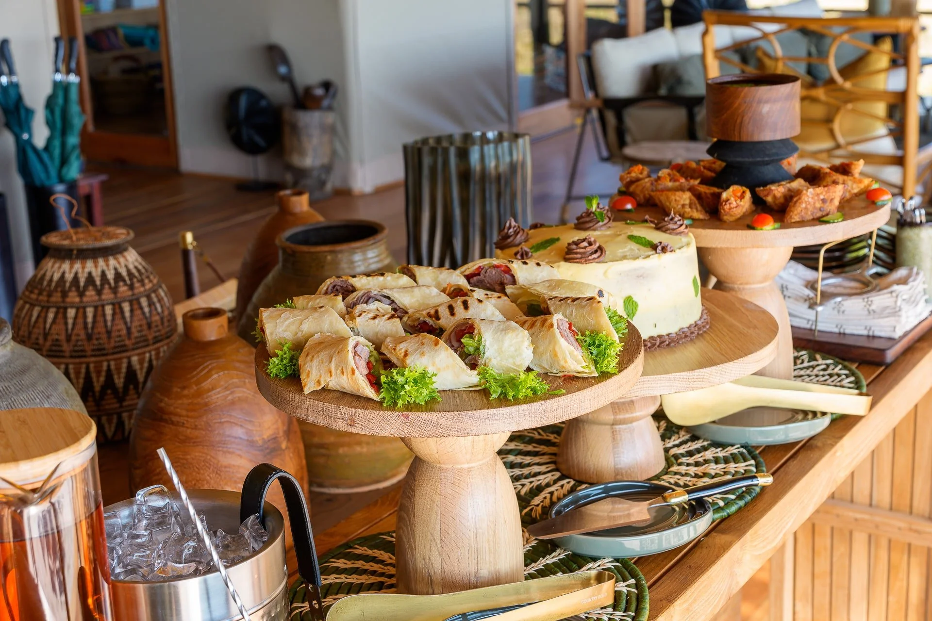 Display of assorted desserts and finger foods on a serving table with wooden stands, including wraps, cake, and savory bites, with ceramic and wicker vases in the background.