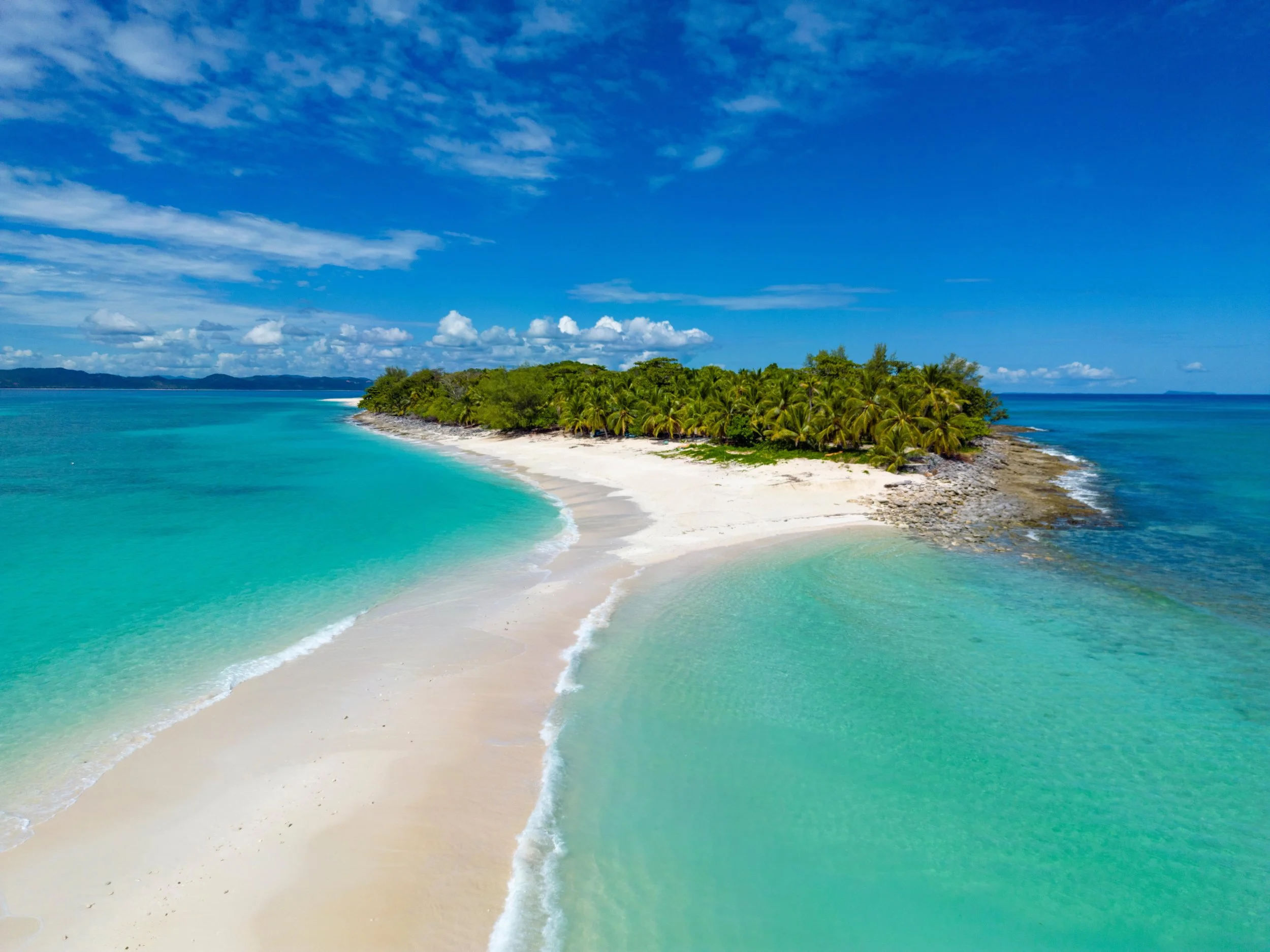 A tropical island with white sandy beaches, turquoise waters, and lush green palm trees under a bright blue sky with some clouds.