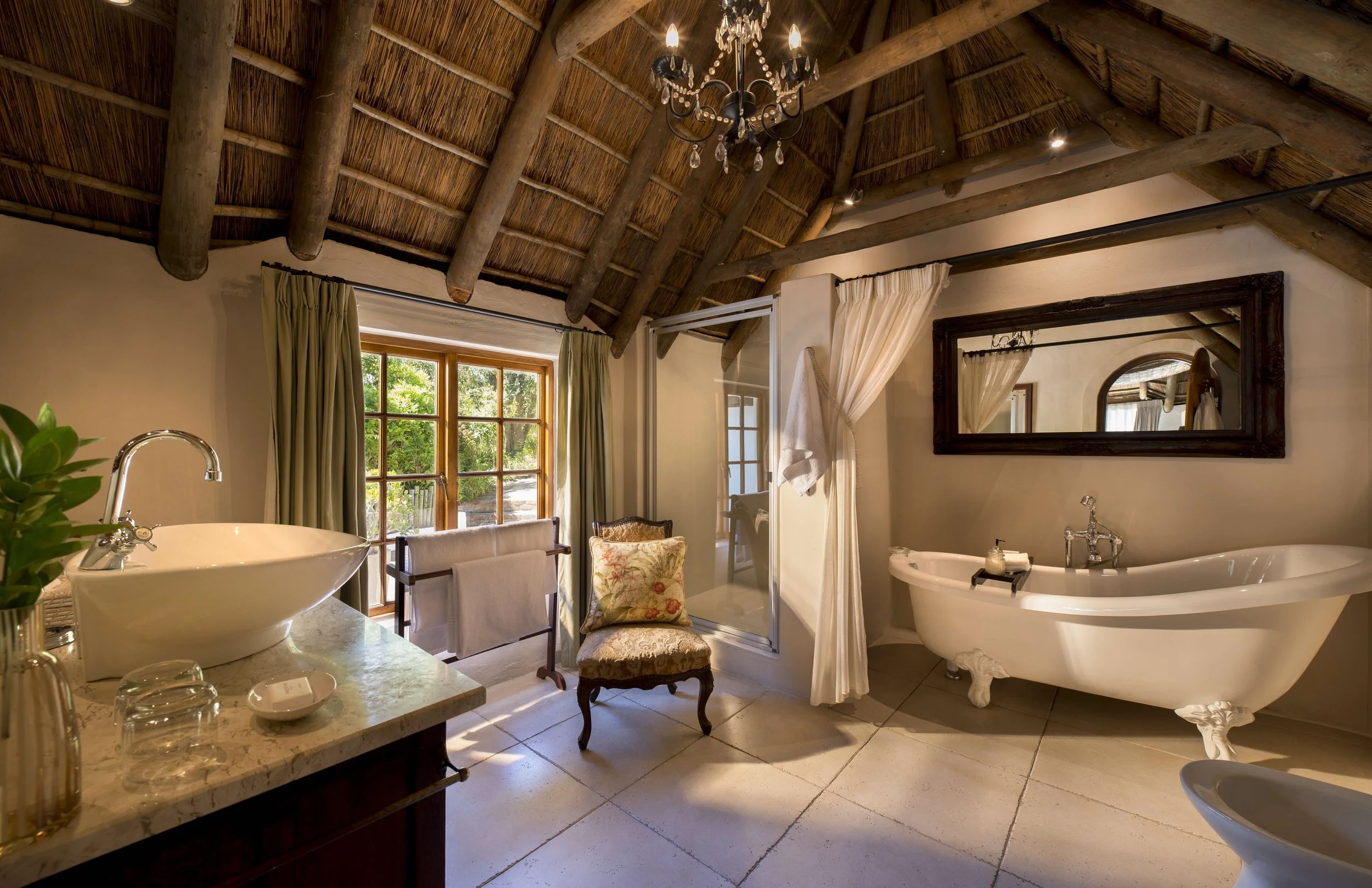 A rustic bathroom with a thatched roof, sunlight coming through a window, a freestanding bathtub, a glass shower enclosure, a mirror, a chair, and various bathroom essentials.