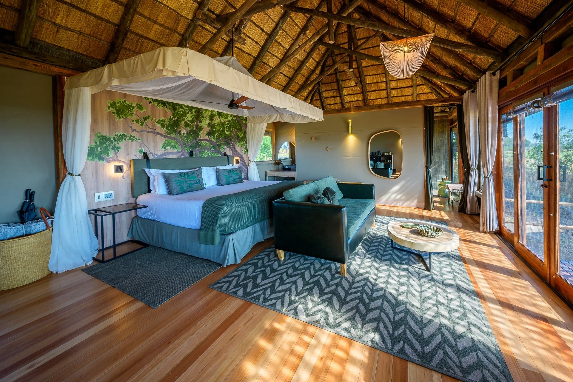 Luxury bedroom with canopy bed, nature-inspired decor, large windows with wooden frames, sunlight streaming in, cozy seating area, patterned rug, and wood-paneled ceiling.