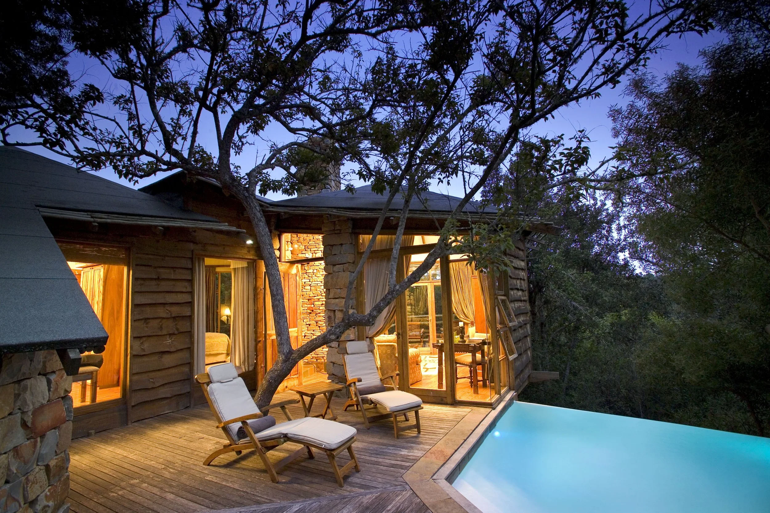 A cozy wooden cabin with large glass windows and a sliding door, illuminated from inside, overlooking a wooden deck with two lounge chairs and an infinity pool, surrounded by trees during twilight.