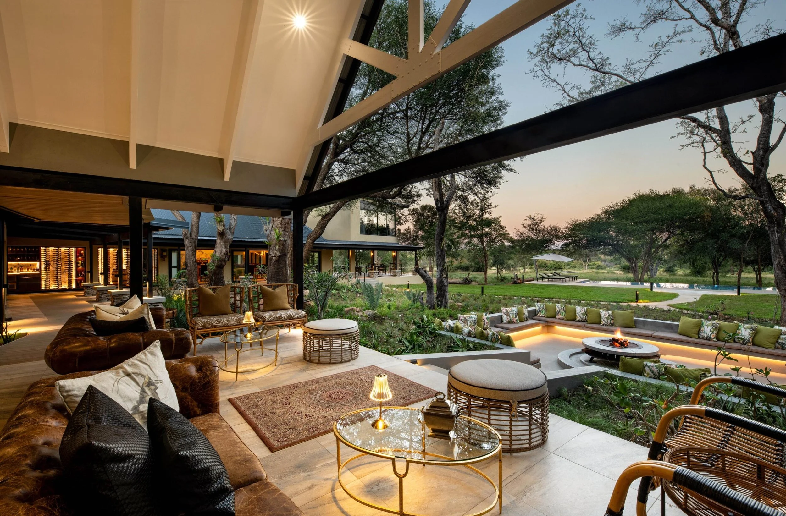 Luxury outdoor patio seating area with leather sofas and chairs, illuminated lanterns, and a fire pit, overlooking a green landscape with trees and a body of water at dusk.