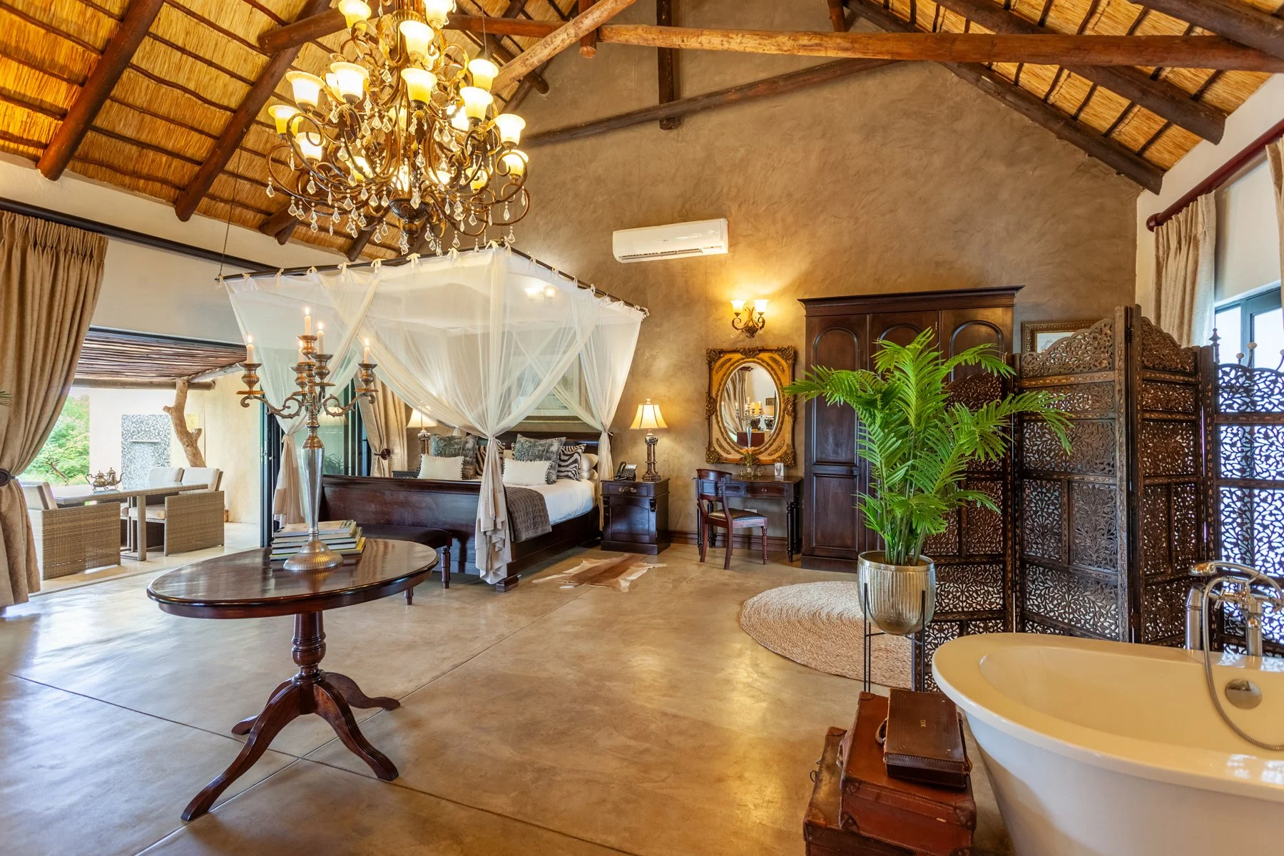 Luxury bedroom with a canopy bed, chandelier, ornate mirror, wooden furniture, large green plant, and a freestanding bathtub in a spacious room with high, thatched ceiling.