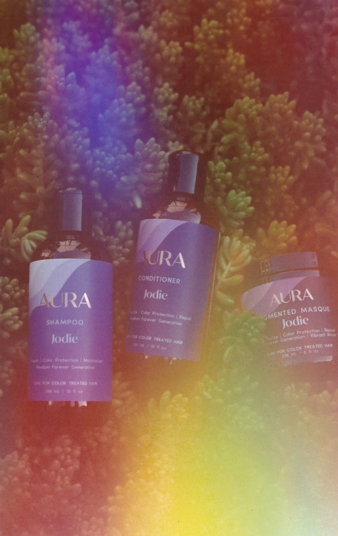 Three bottles of Aura hair care products—shampoo, conditioner, and a mask—placed on a background of green succulent plants. The bottles are purple with white labels.
