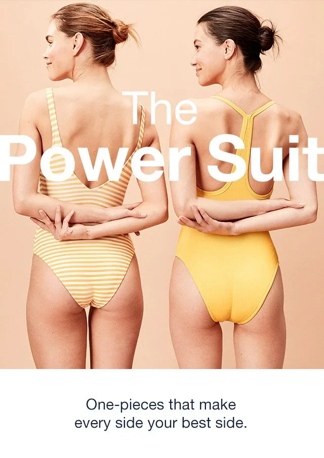 Two women standing with their backs to the camera, one in a yellow and white striped power suit and the other in a solid yellow power suit. Both are smiling slightly and holding their arms behind their backs. Text overlay reads "The Power Suit" and b