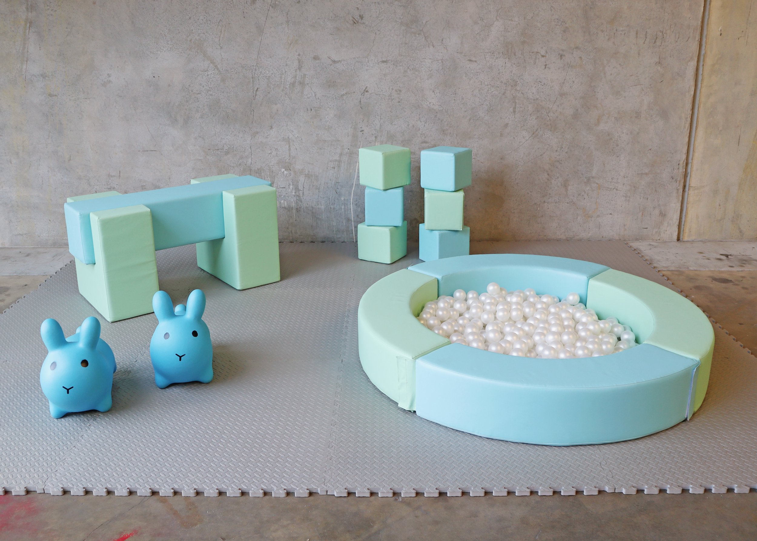 Soft pastel-colored play area with a ball pit filled with white balls, two bunny-shaped toys, a small foam bench, and foam blocks on a gray rubber mat against a concrete wall background.