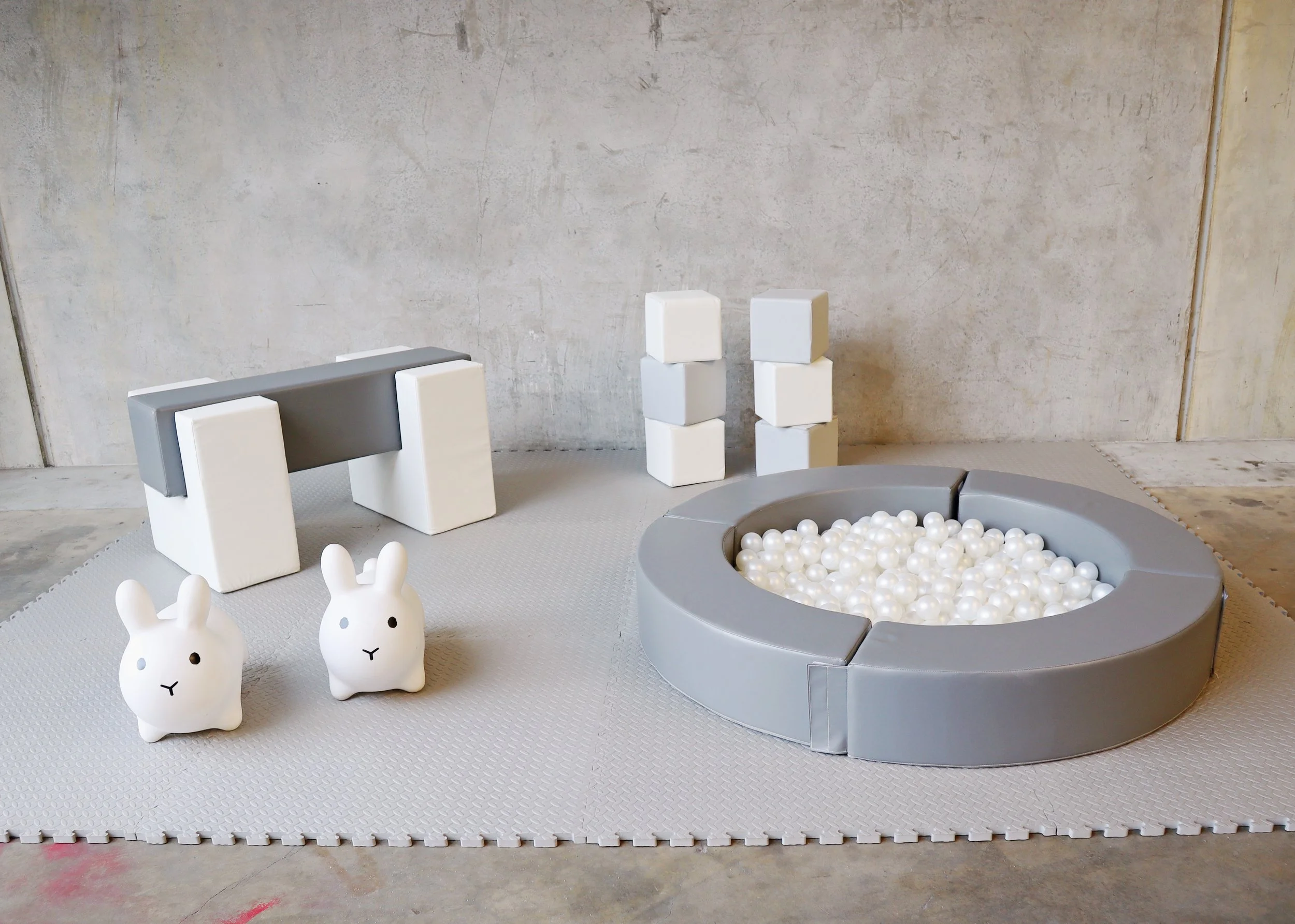 Play area with soft mats, two bunny-shaped toys, white boulders stacked in two columns, and a round ball pit filled with white balls, against a plain wall.