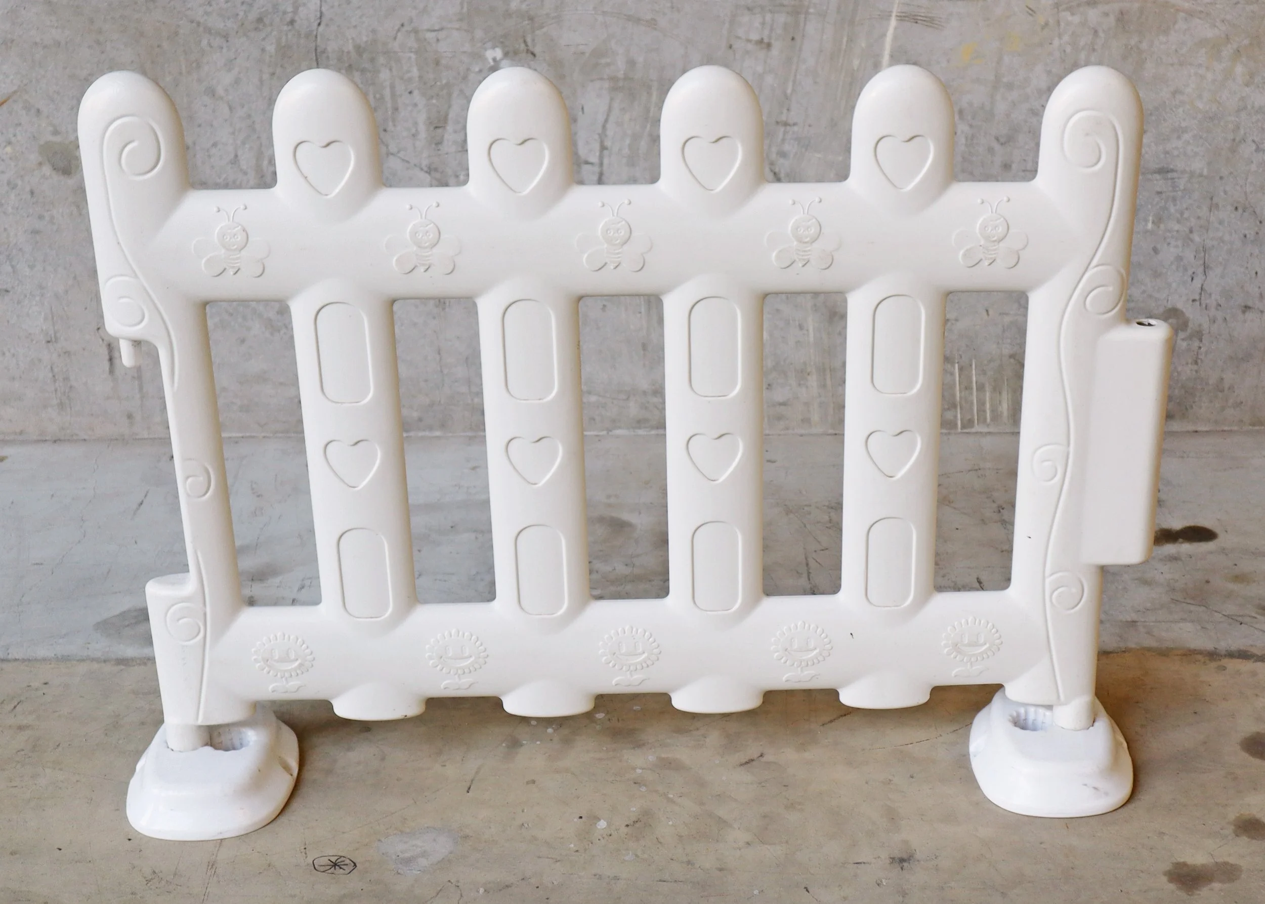 White plastic safety barrier with heart and bee motifs, placed on a concrete surface against a concrete wall.