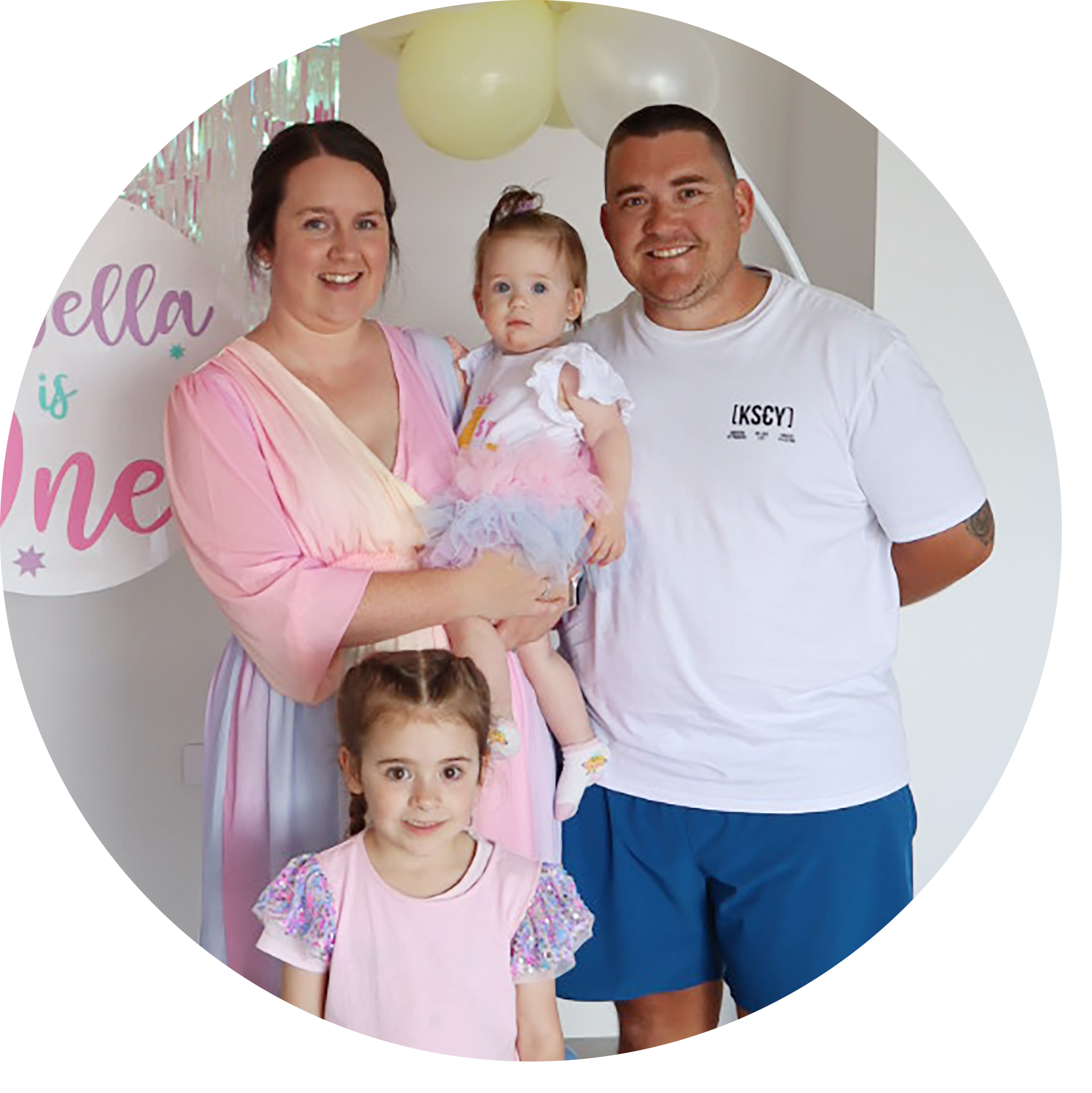 Family standing together at a celebration, with a woman holding a baby girl in a tutu, a young girl in front wearing a pink shirt with sparkly sleeves, and a man with short hair and a tattoo on his arm, all smiling.