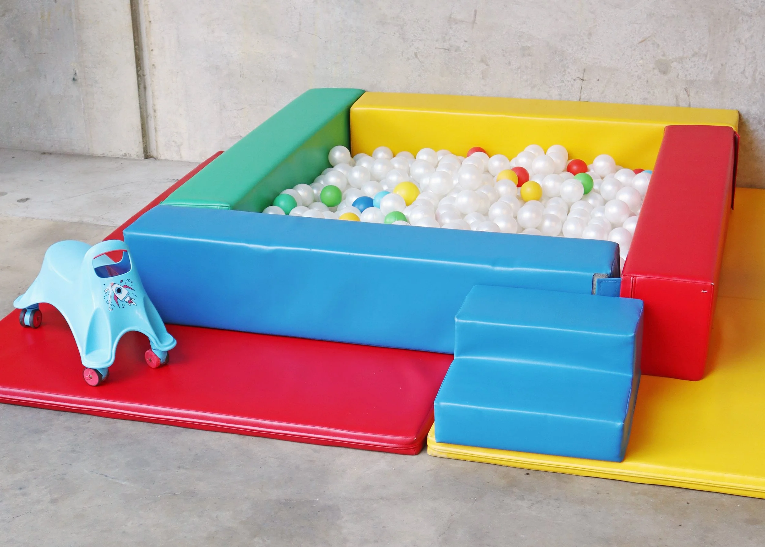 Colorful children's ball pit with a red, green, yellow, and blue padded walls filled with white, green, yellow, blue, and red plastic balls, arranged on a red and yellow padded floor with a small blue plastic slide and a blue ride-on toy in front.