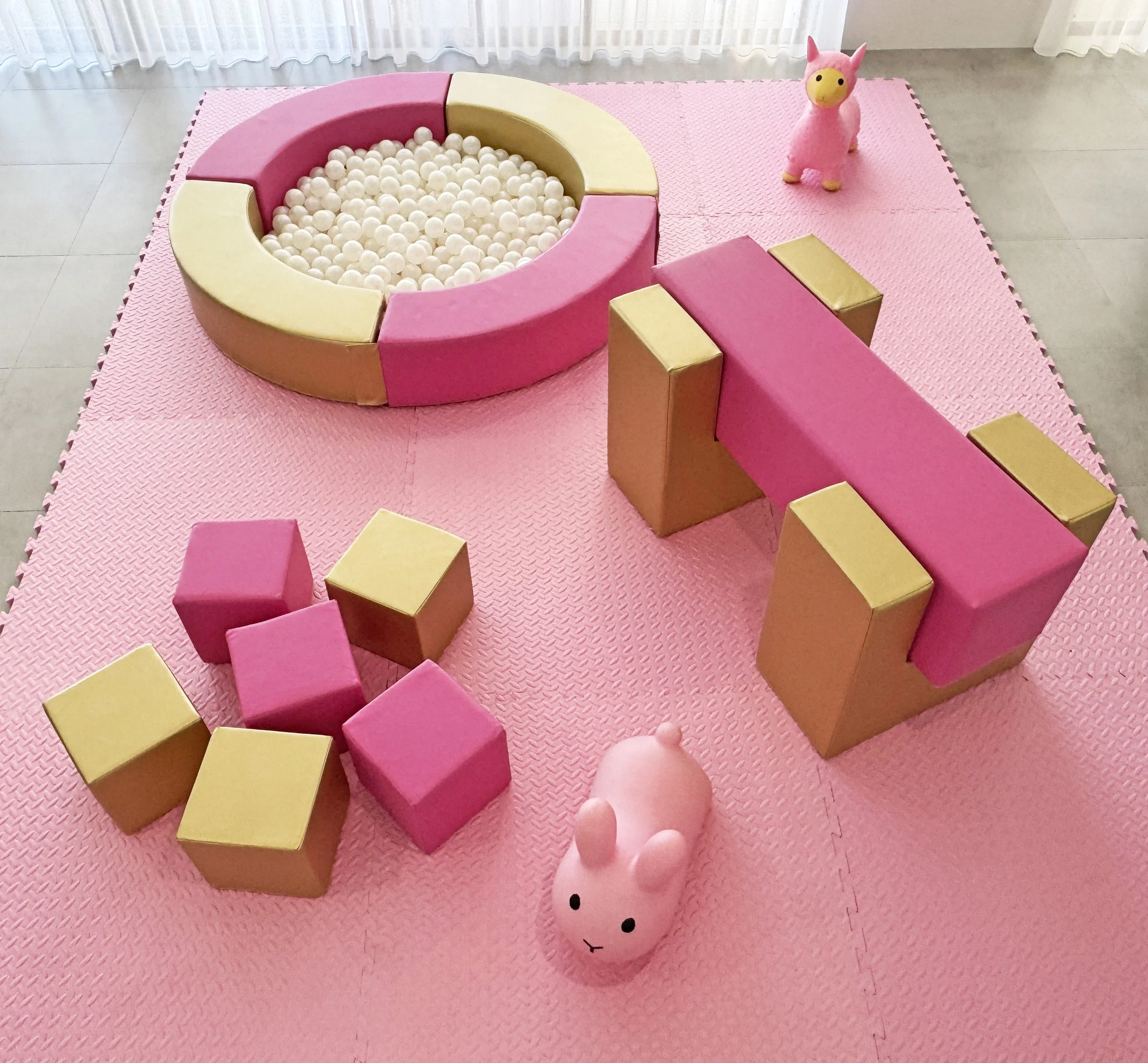 Children's play area with pink and gold foam blocks, a ball pit with white balls, a plush pink bunny toy, and a pink plush llama toy on a pink foam mat.