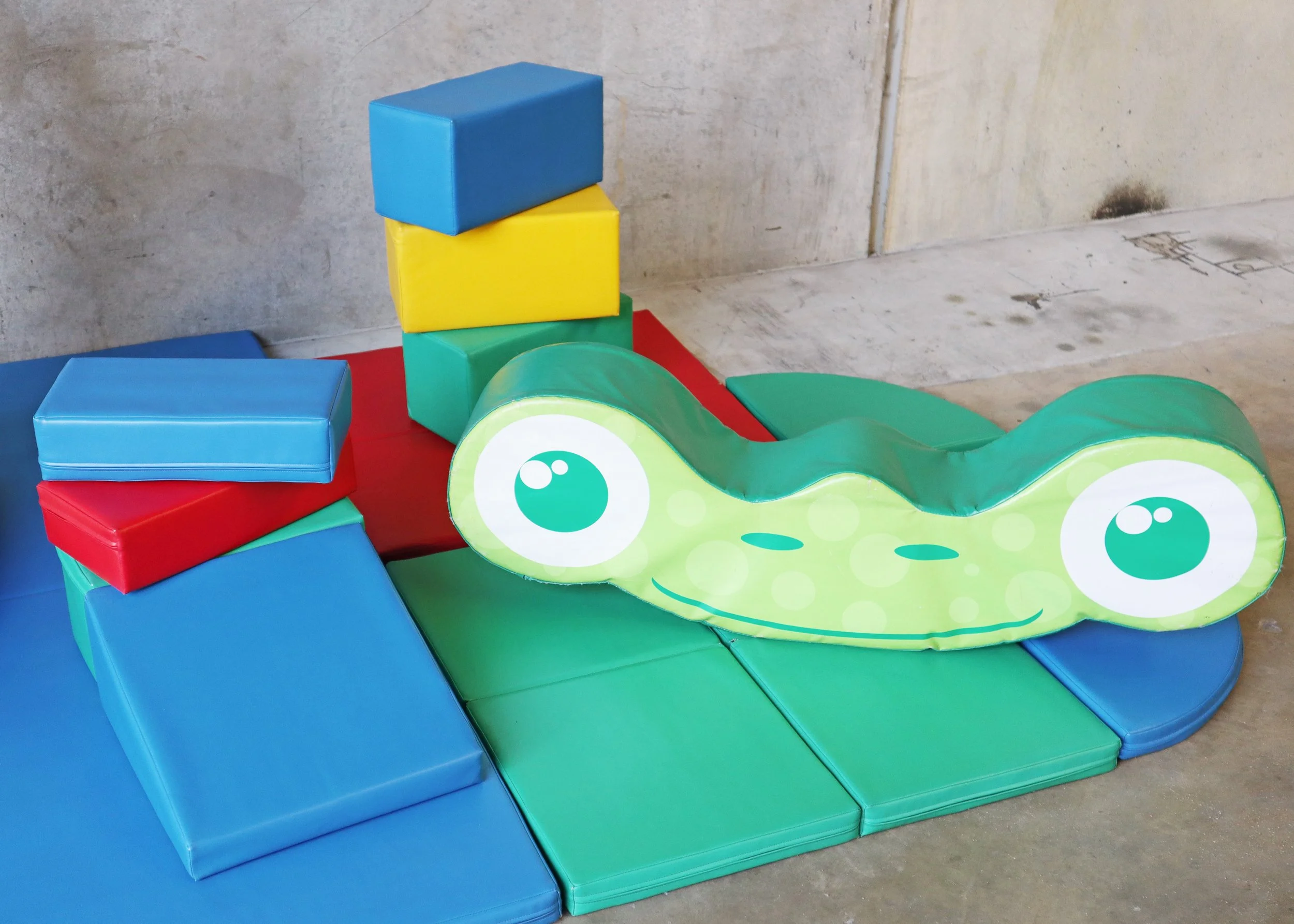 Colorful foam play area with soft blocks and a green alligator-shaped cushion with large cartoon eyes, set against a concrete wall.