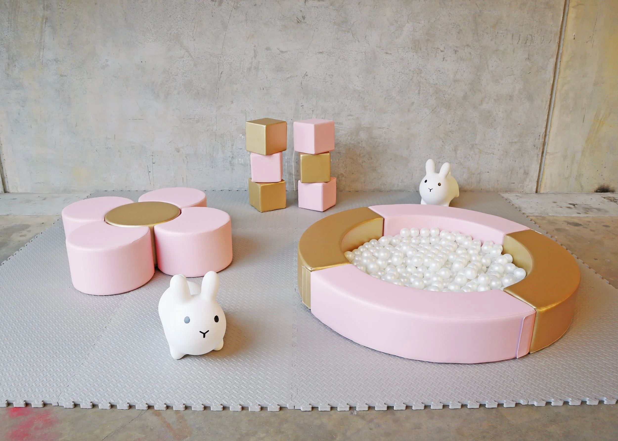 A children's play area with pink and gold foam furniture, including a large circular ball pit and smaller stools, accompanied by two white rabbit toys and small pink and gold stacking blocks, all on a soft gray mat against a concrete wall.