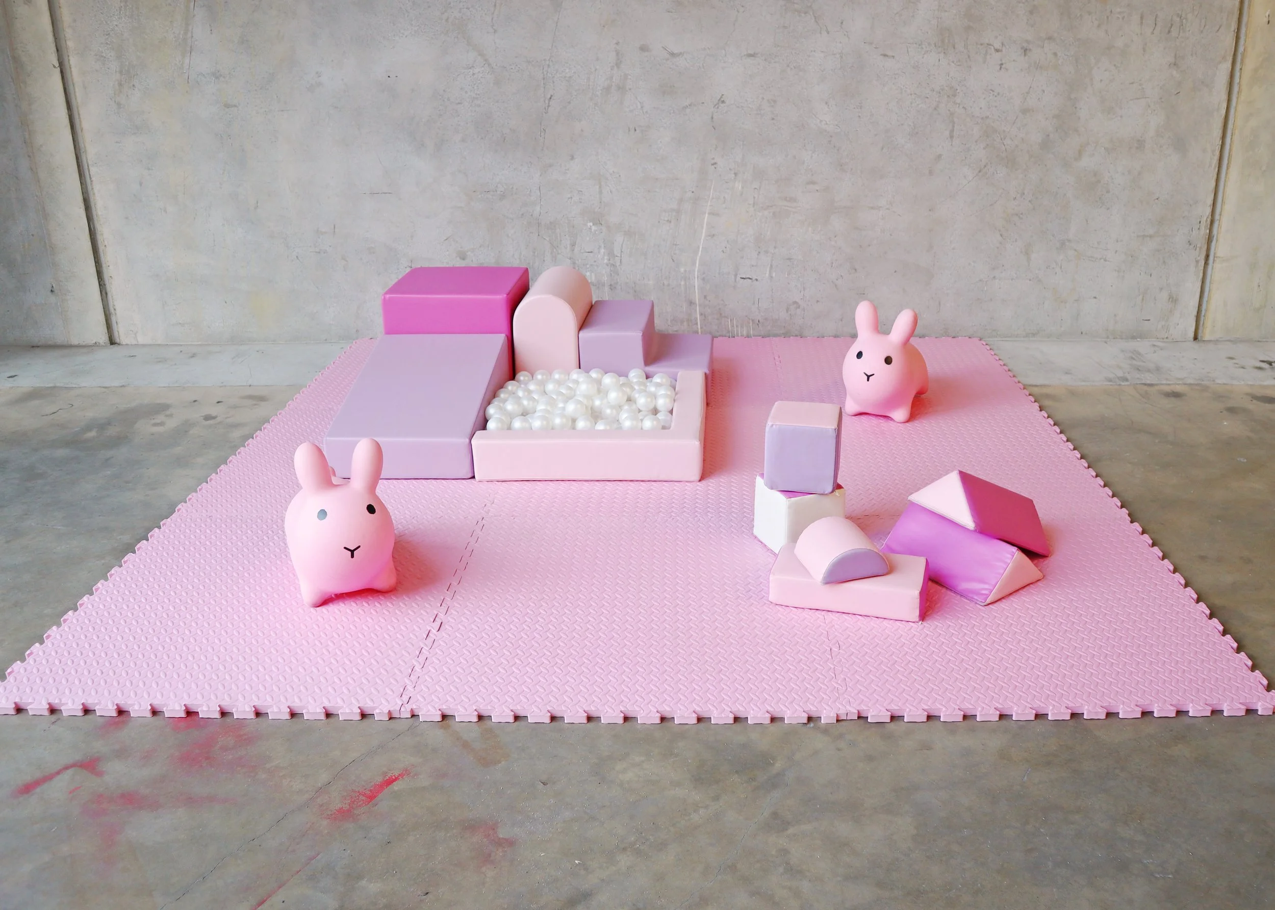 Pink foam play mat with pastel-colored soft blocks, a small pink bunny figurine, and a white bunny figurine, set against a plain concrete wall.
