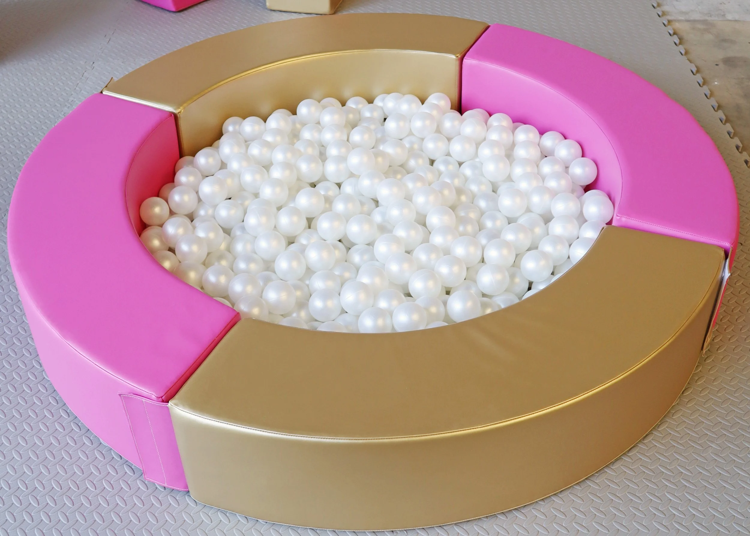 A round pink, gold, and beige ball pit filled with white plastic balls, set on a textured floor.