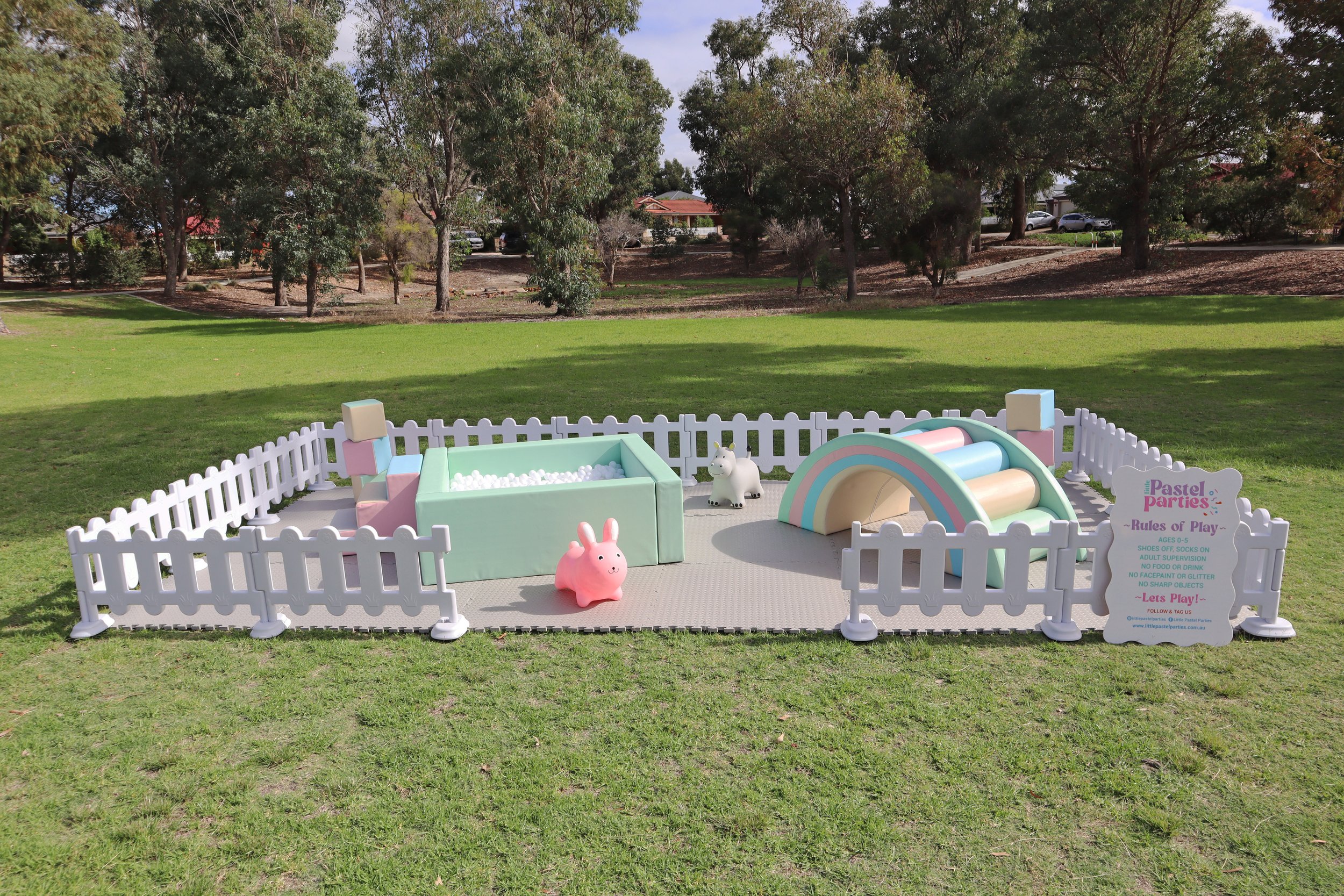 Small fenced play area with pastel-colored toys, including a bunny-shaped ride, ball pit, rainbow tunnel, and a unicorn figurine, set on a grassy field with trees in the background.