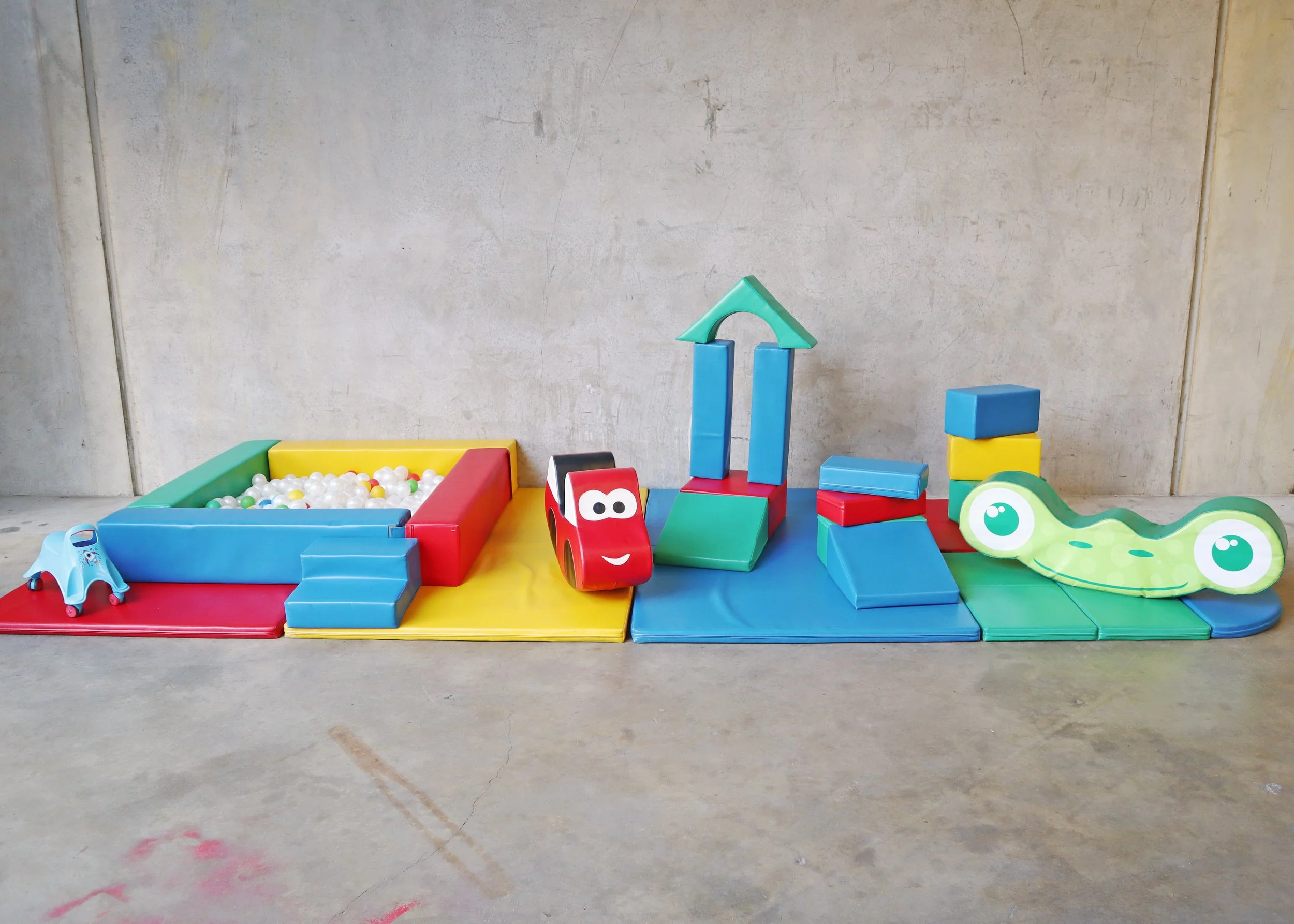 Colorful children's foam play set with ball pit, toy car, foam blocks, and a smiling frog figure against a concrete wall.