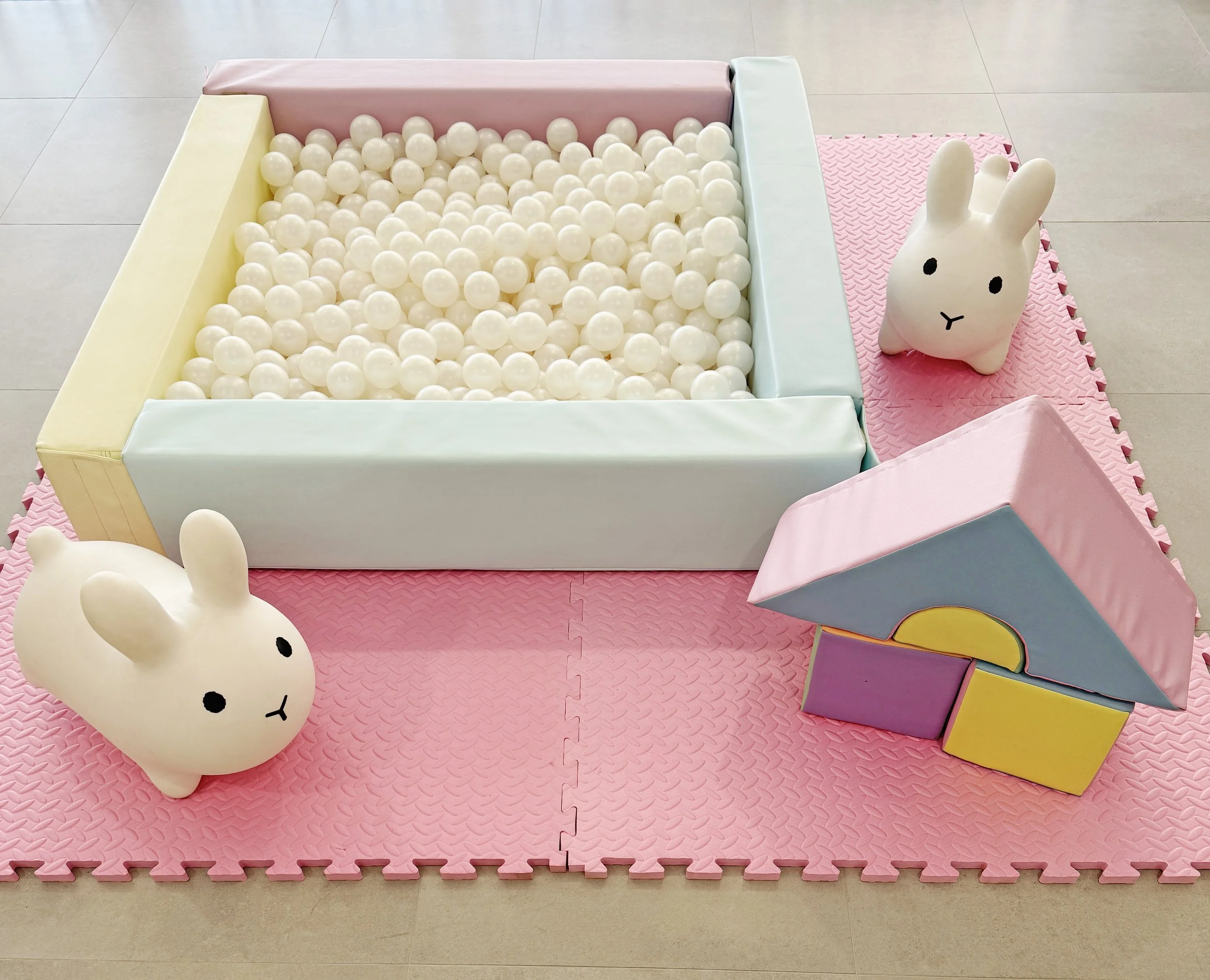 Children's play area with a soft pink foam mat, a white ball pit with pastel-colored padded walls, two white rabbit-shaped plush toys, and a colorful foam block structure resembling a small house.