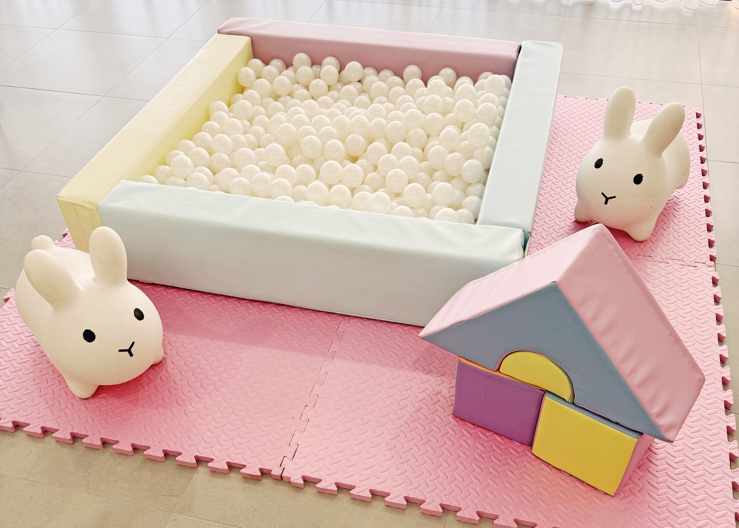 Children's play area with a pink foam mat, a ball pit with white balls, and two bunny-shaped soft toys.