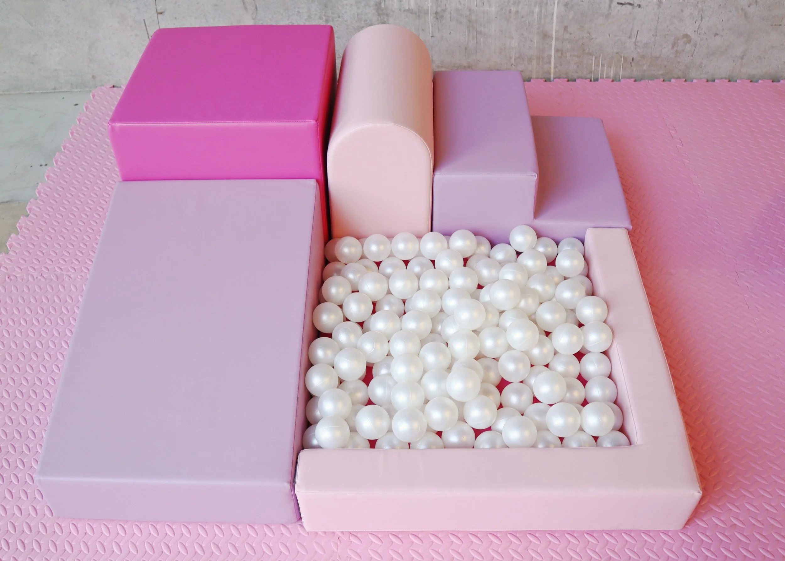 Colorful padded playroom corner with pastel pink, purple, and cream blocks surrounding a ball pit filled with white plastic balls, on a pink textured rubber mat.
