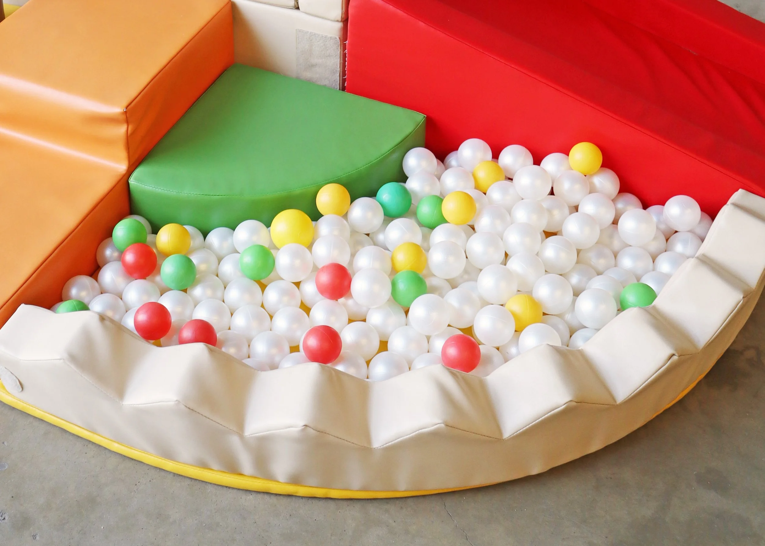 Children's ball pit with white, yellow, green, and red plastic balls, surrounded by padded orange, red, green, and white walls.