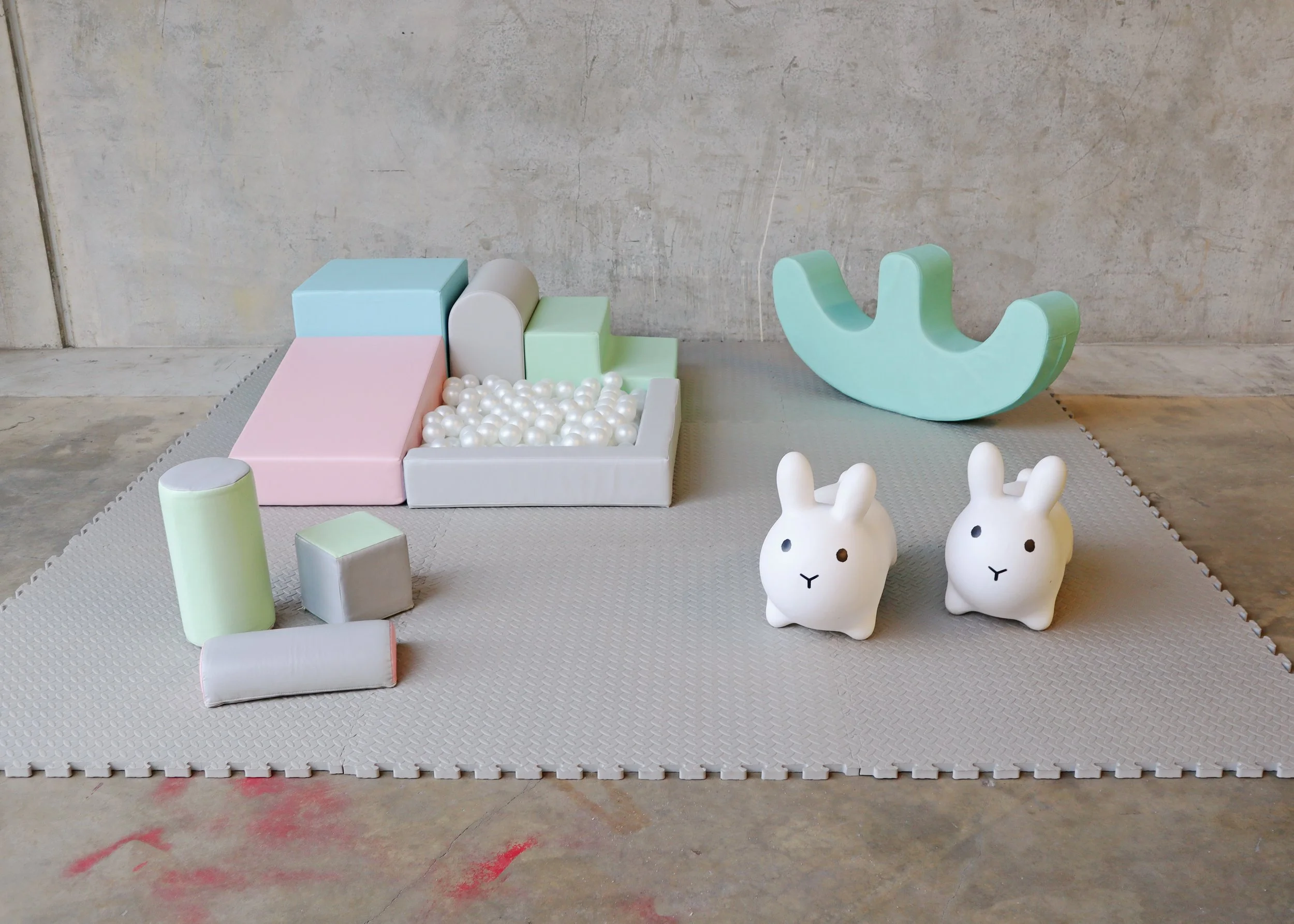 Colorful children's toys arranged on a soft play mat against a concrete wall, including geometric blocks, a ball pit filled with white plastic balls, two white rabbit-shaped figurines, and a mint green crescent-shaped rocker.