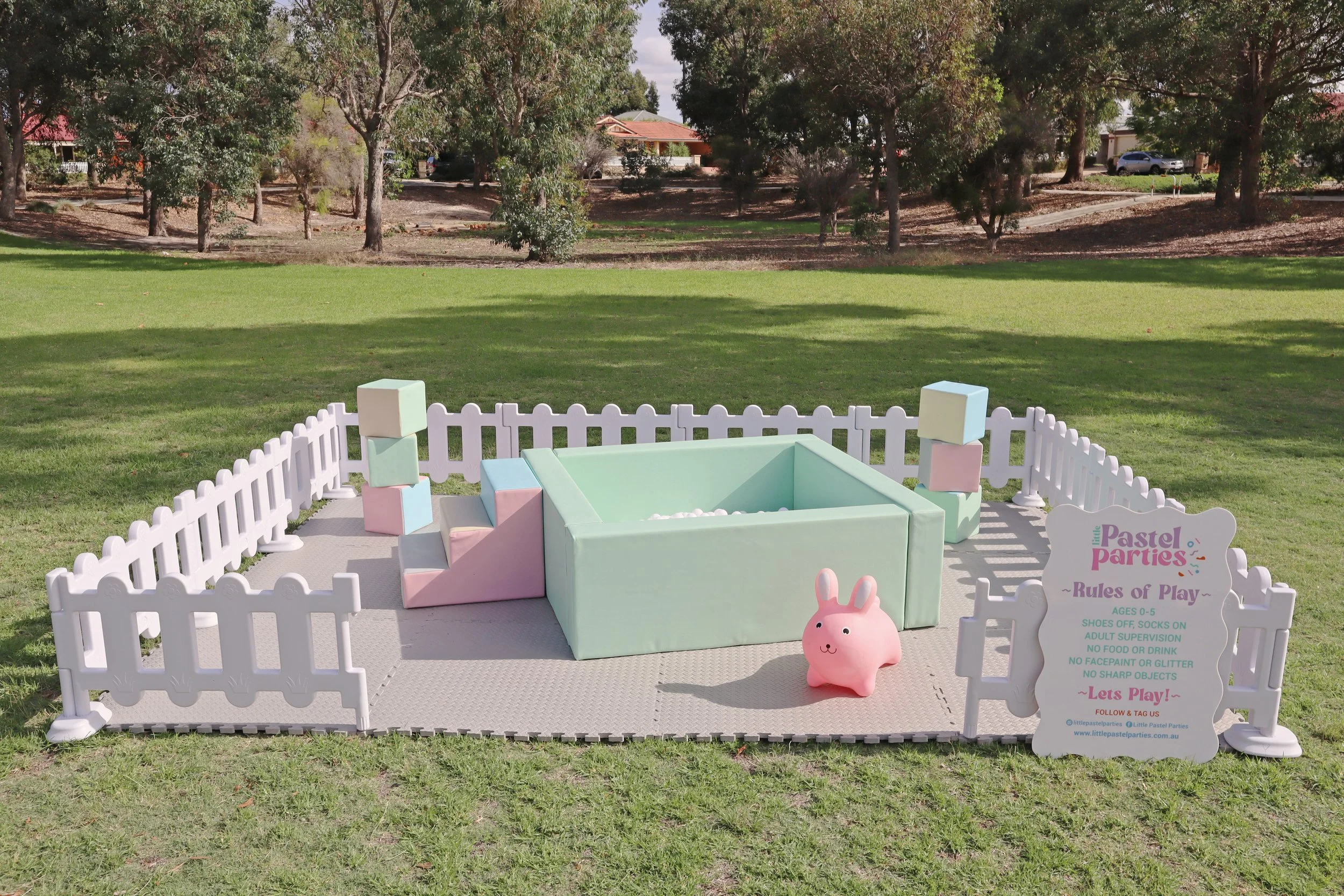 A pastel-themed outdoor play area for children aged 0-5, enclosed by a white picket fence. Inside, there are soft pastel-colored blocks and a green soft play square with white pebbles. A pink bunny-shaped toy is in front. A signboard lists the rules of play, including no shoes, socks on, adult supervision, no food or drink, no face paint or glitter, and no sharp objects.