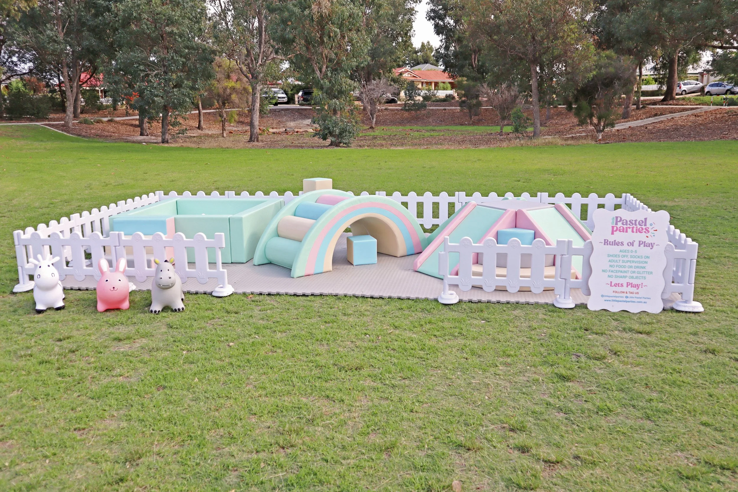 Colorful pastel playground enclosed by a white picket fence, featuring a rainbow tunnel, a pyramid climbing structure, and bunny-shaped inflatable toys on grass.