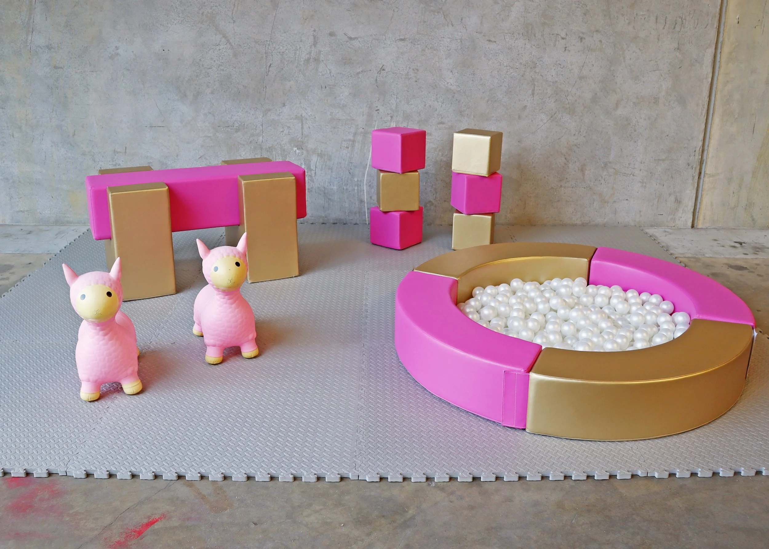 Colorful play area with pink and gold foam blocks, two pink llama toys, and a ball pit filled with white plastic balls, set against a concrete wall.