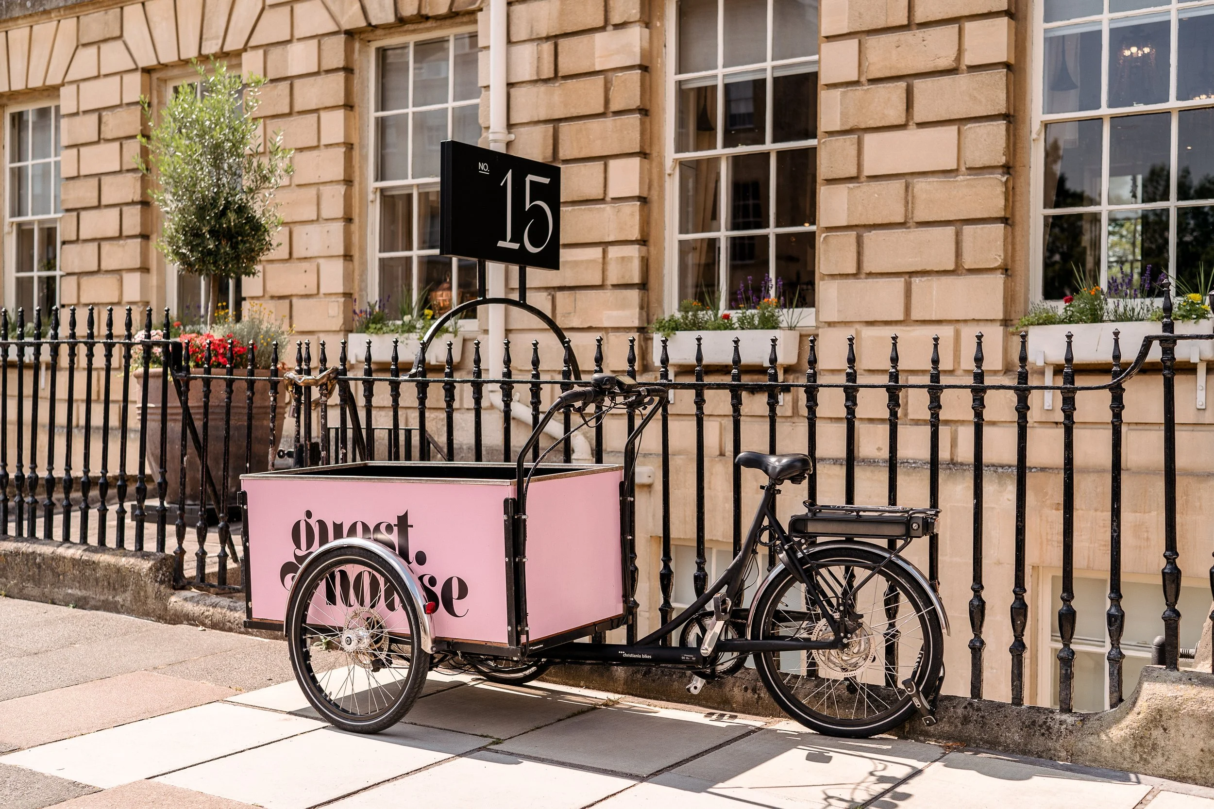 A bicycle with a pink cargo box that says 'guest. love' parked in front of a stone building with white-framed windows and flower boxes. A black fence surrounds the area.