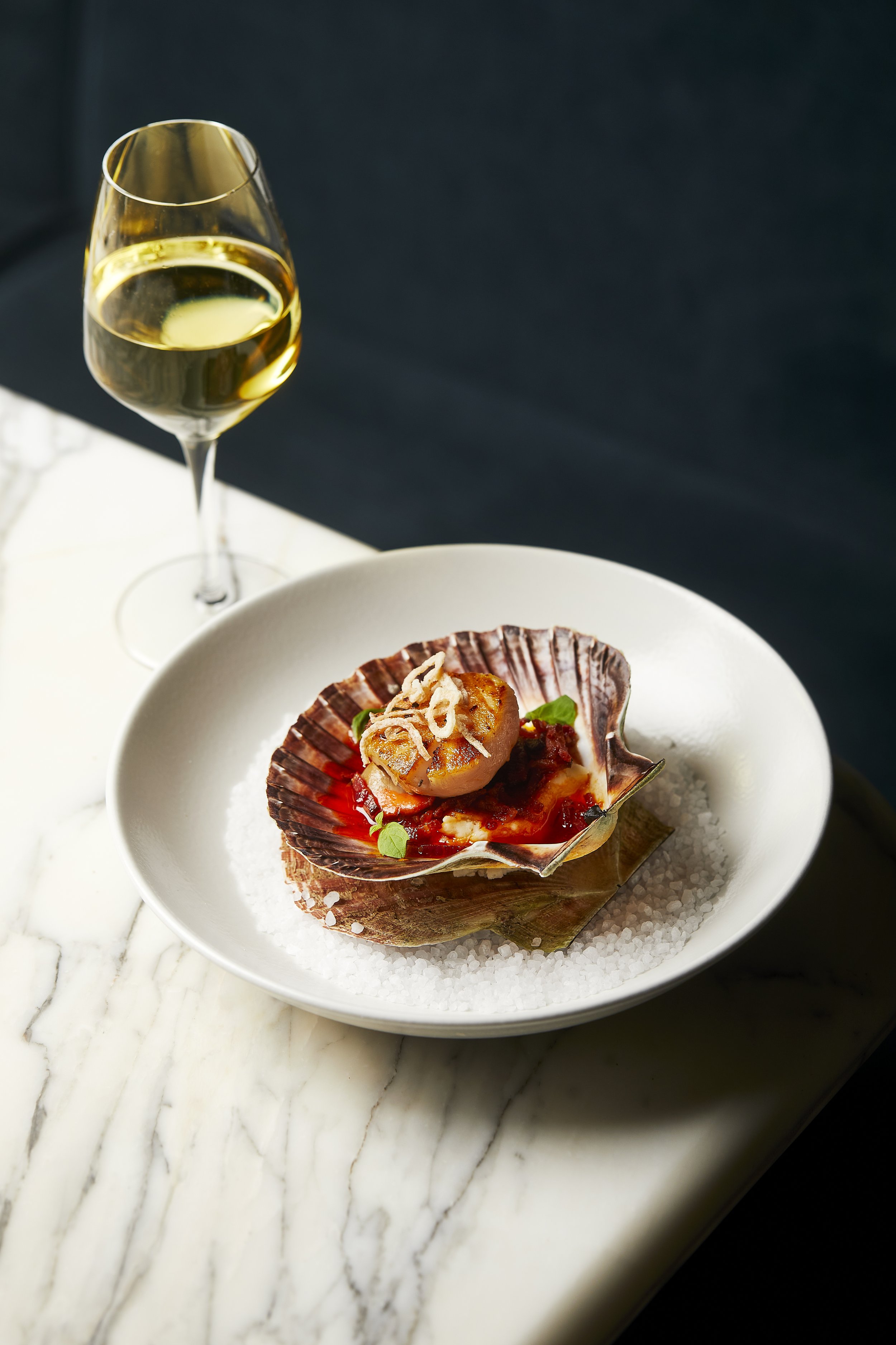 A glass of white wine next to a plate with a seafood dish served in a shell, with a bed of salt surrounding the shell on a white marble surface.