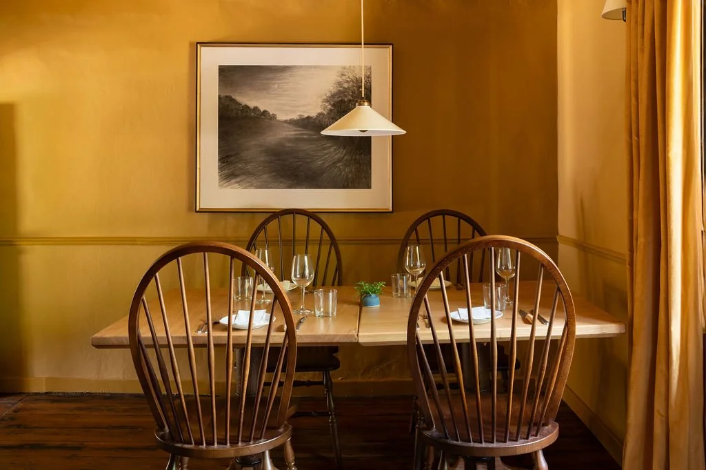 A wooden dining table set for four with wine glasses, water glasses, and plates with napkins in a yellow-walled room. A framed landscape painting hangs on the wall, and a white pendant light hangs above the table. Golden curtains are on the right.