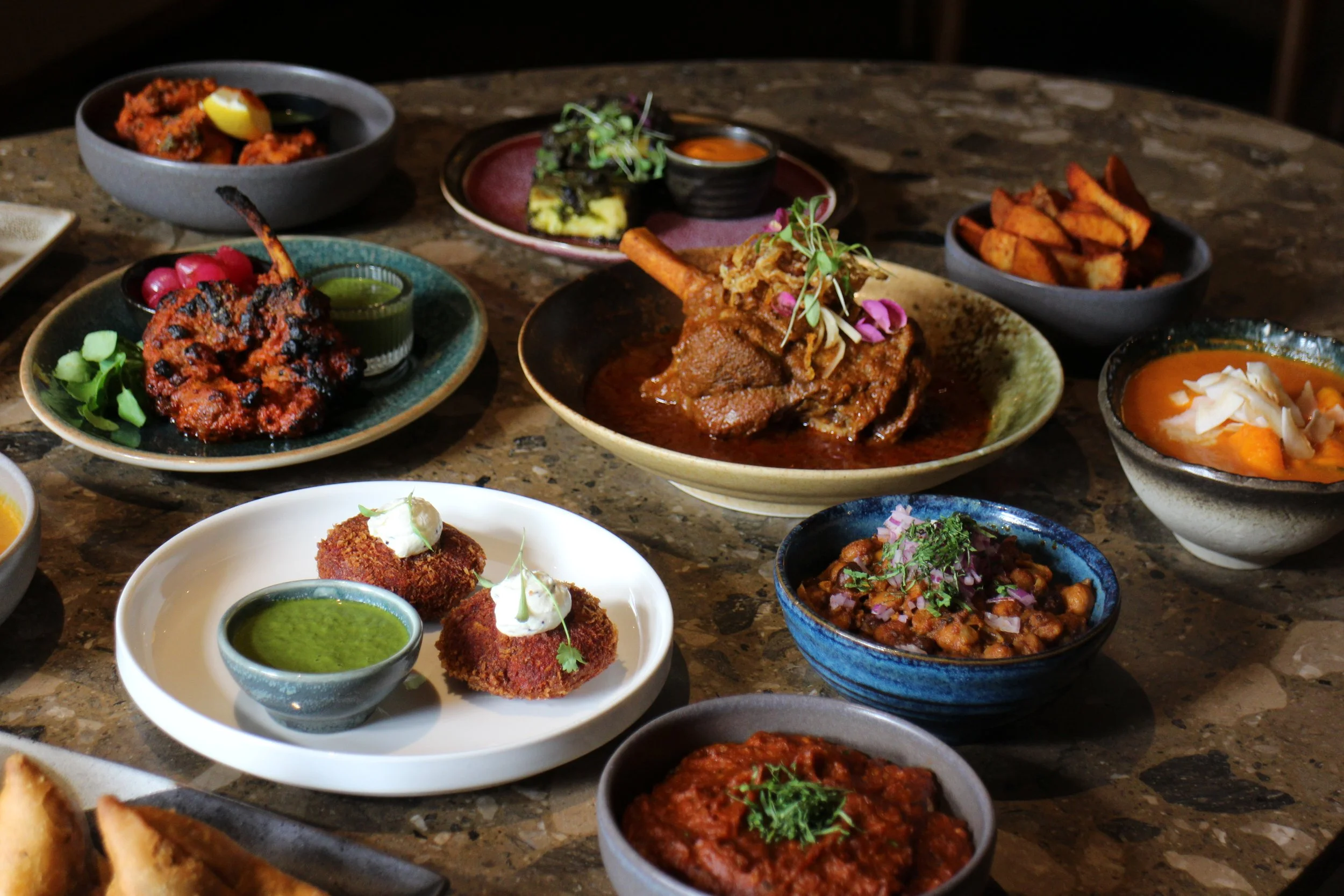 A variety of Indian dishes served on a dark marble table, including curries, fried snacks, and chutneys. Made by Kokum a leading upmarket Indian Restaurant in South London one of Amy Williams Consultancy's clients