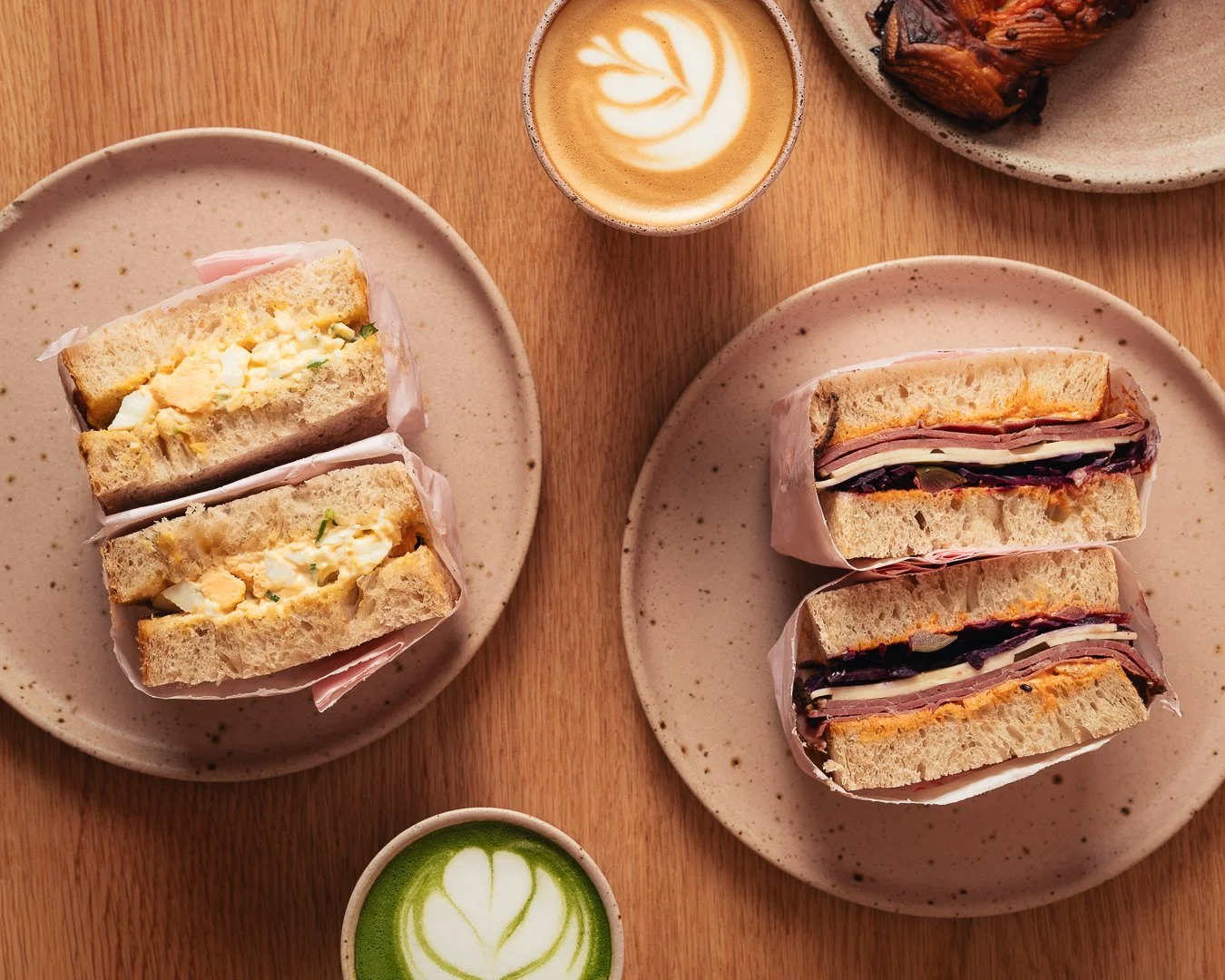 Two plates of sandwiches, a cup of coffee with latte art, a small green matcha latte, and a bowl of chicken wings on a wooden table.