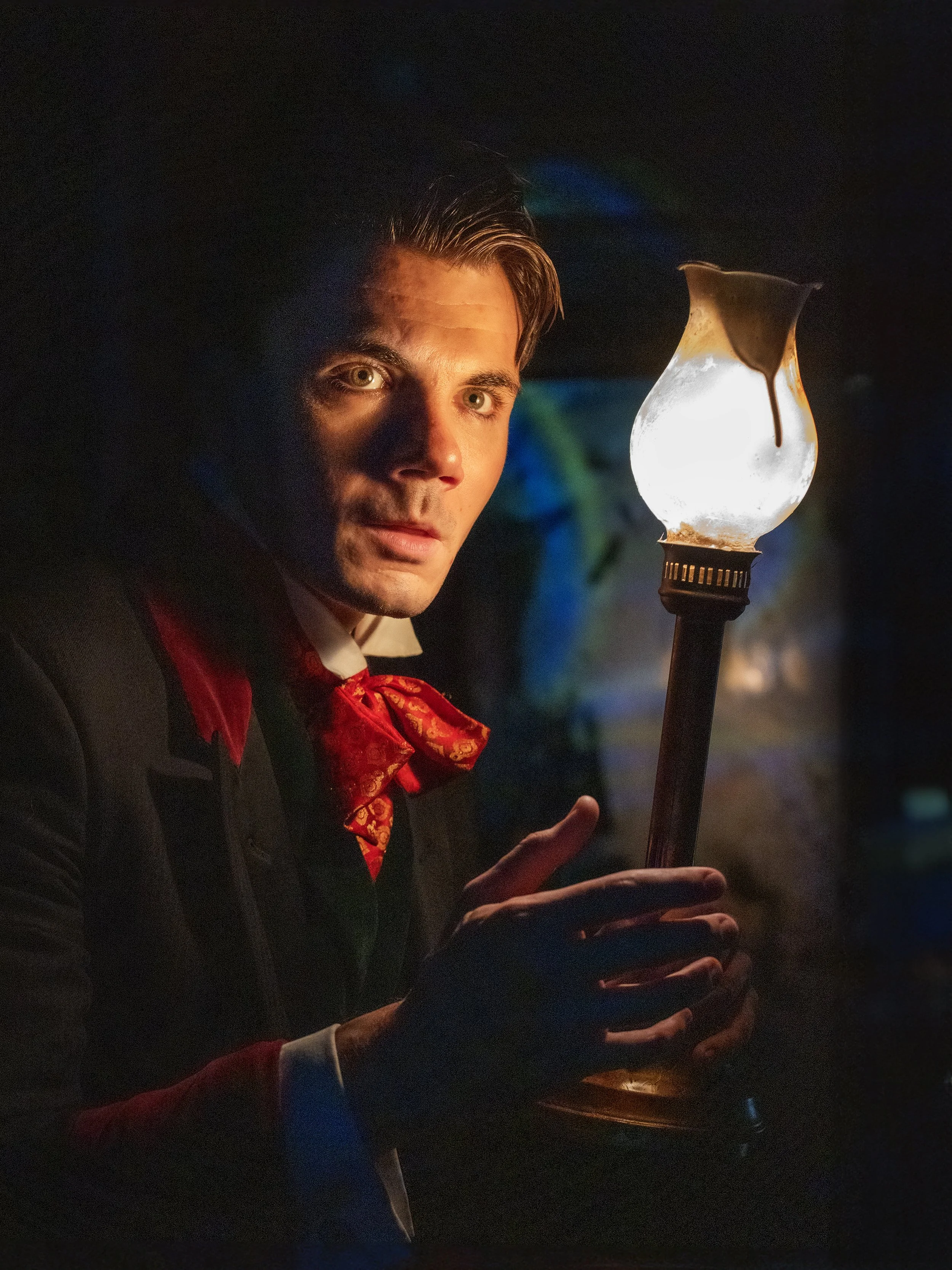 A man in formal attire with a red bow tie holding a glass oil lamp in a dark setting, illuminated by the lamp's light.