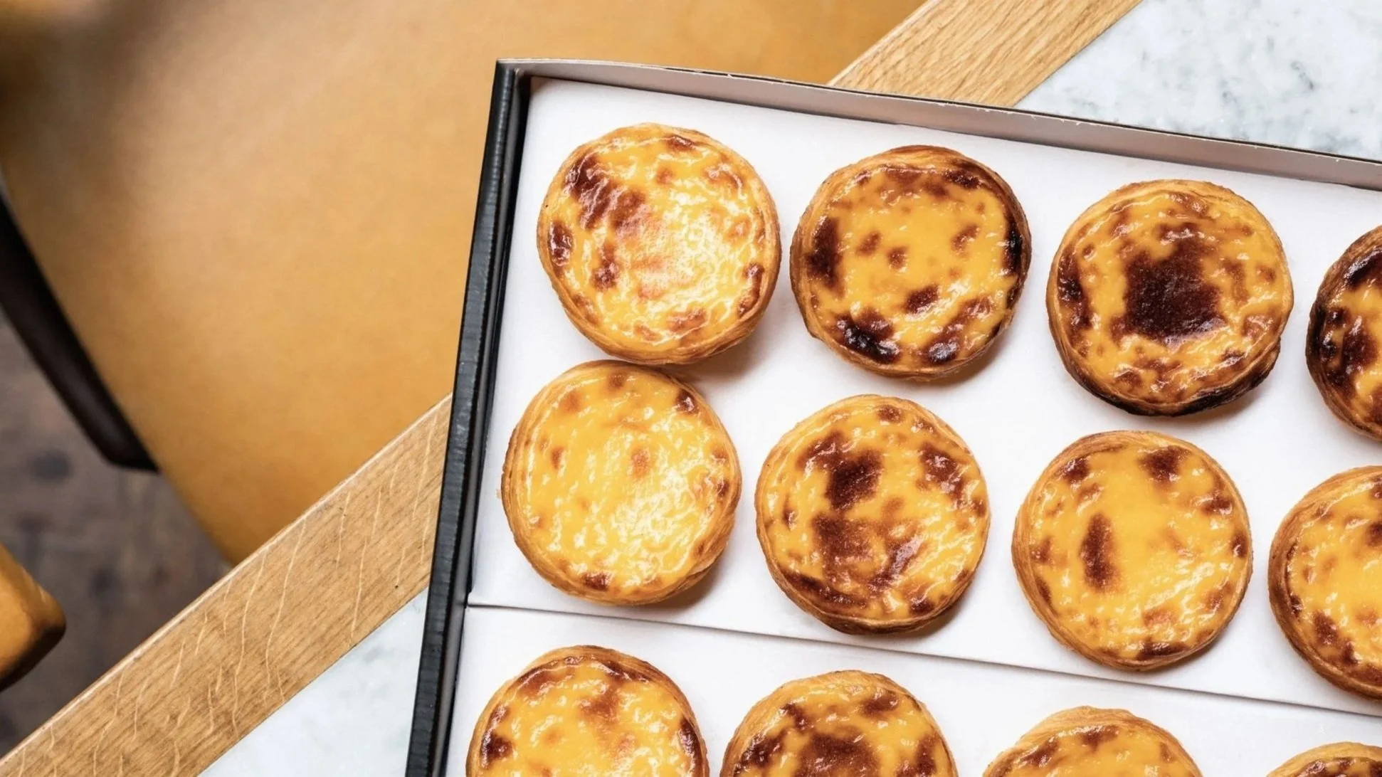 Tray of fresh Portuguese egg tarts with golden crusts and caramelized tops on a wooden table. Pasteis de Nata by Santa Nata