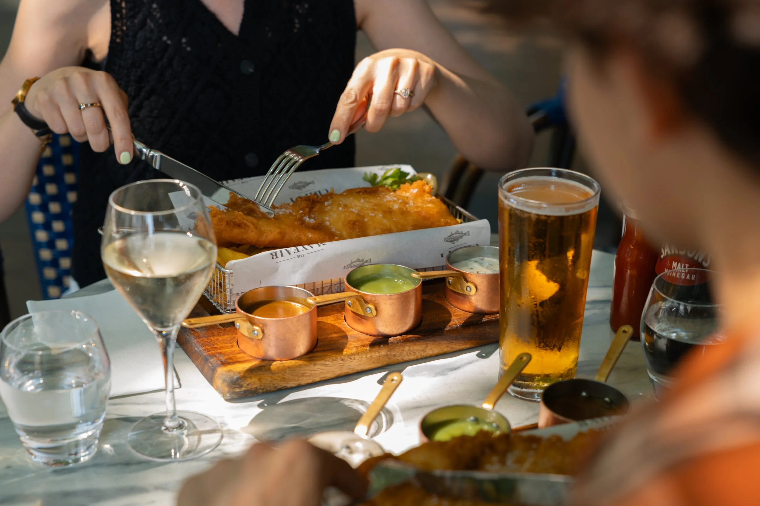 A person with painted nails and rings is cutting a piece of fried fish on a plate at a dining table. The table has various beverages, including beer and water, and multiple small bowls of dipping sauces or condiments.