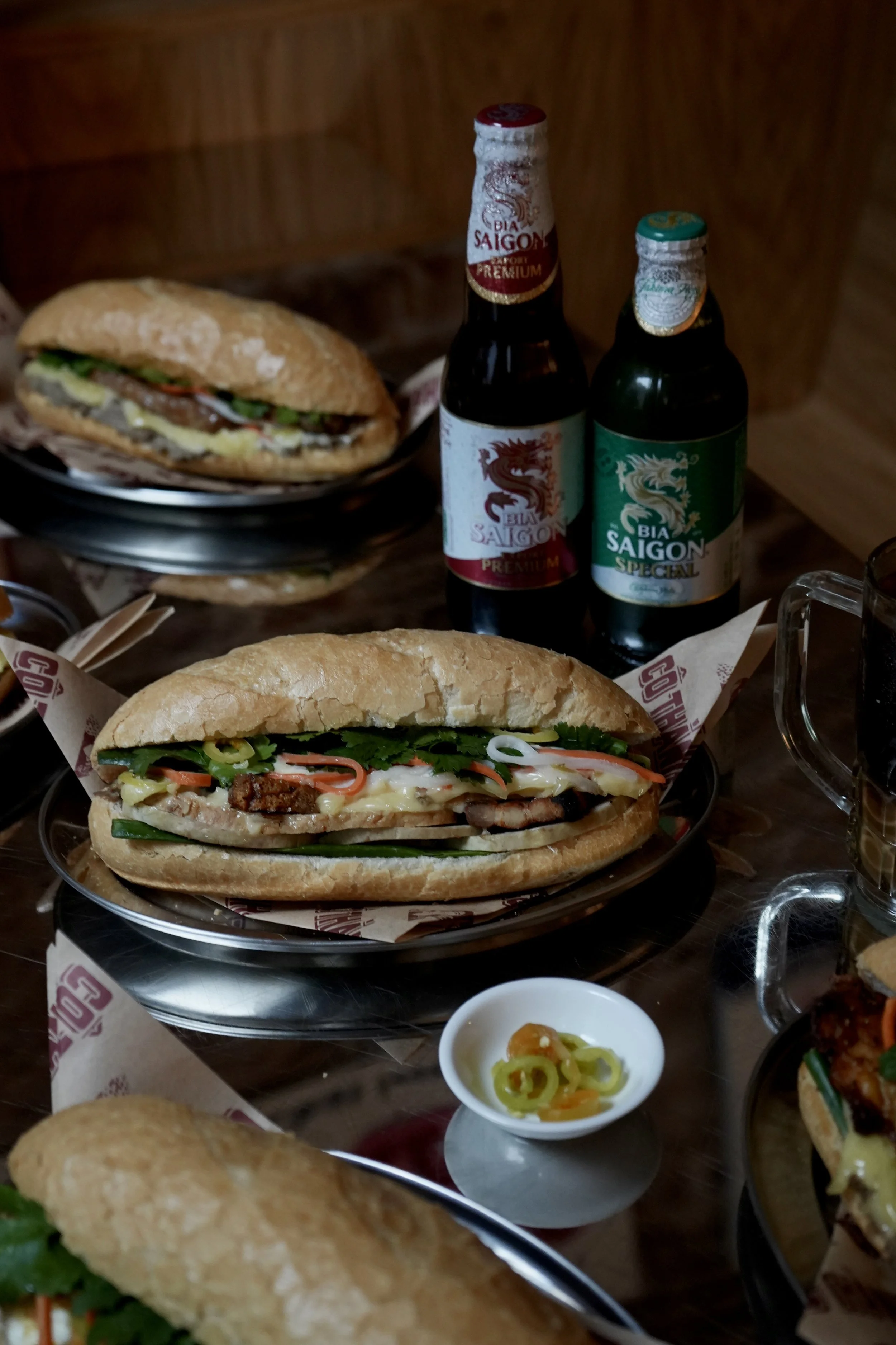 Two Vietnamese banh mi sandwiches with various fillings on black plates, accompanied by bottles of Saigon beer and a small dish of sliced green chili peppers with yellow peppers. By Co Thanh Covent Garden Vietnamese Restaurant an AWC PR Client