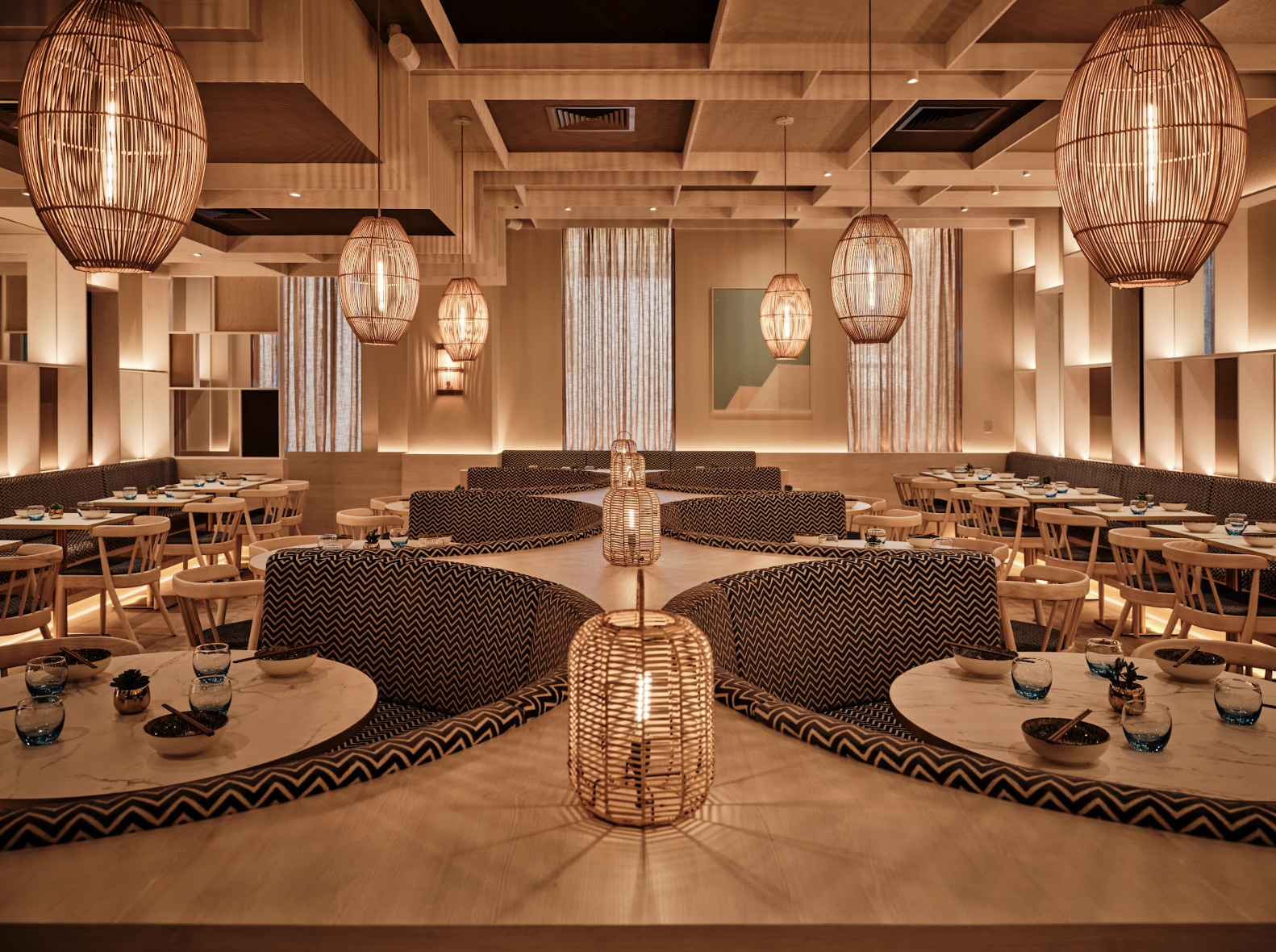 Decorative restaurant dining room with warm lighting, wicker pendant lights, and a mix of round and rectangular tables set with bowls, glasses, and chopsticks.