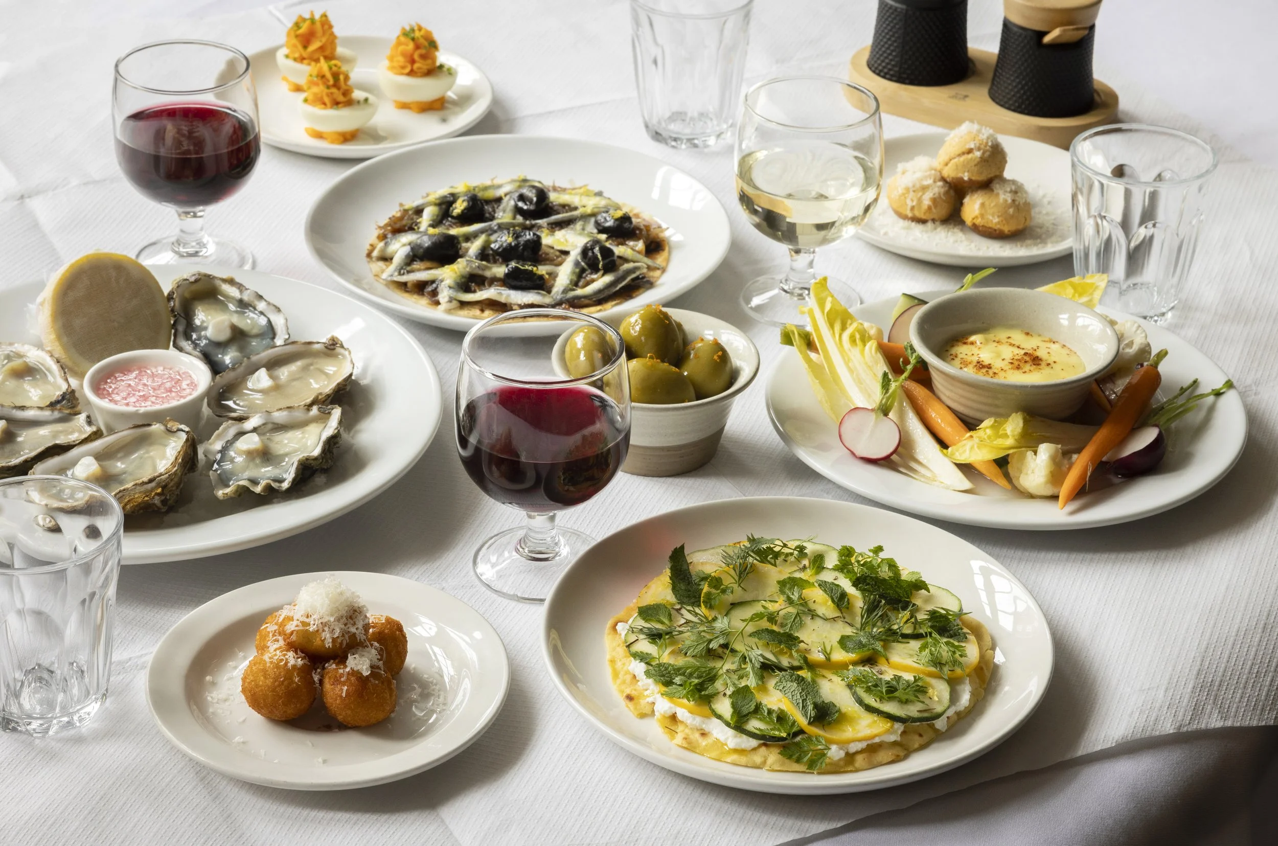 A variety of appetizers and drinks on a white tablecloth, including oysters, a pizza topped with herbs and vegetables, a plate of stuffed eggs, olives, a vegetable salad, and glasses of red and white wine.