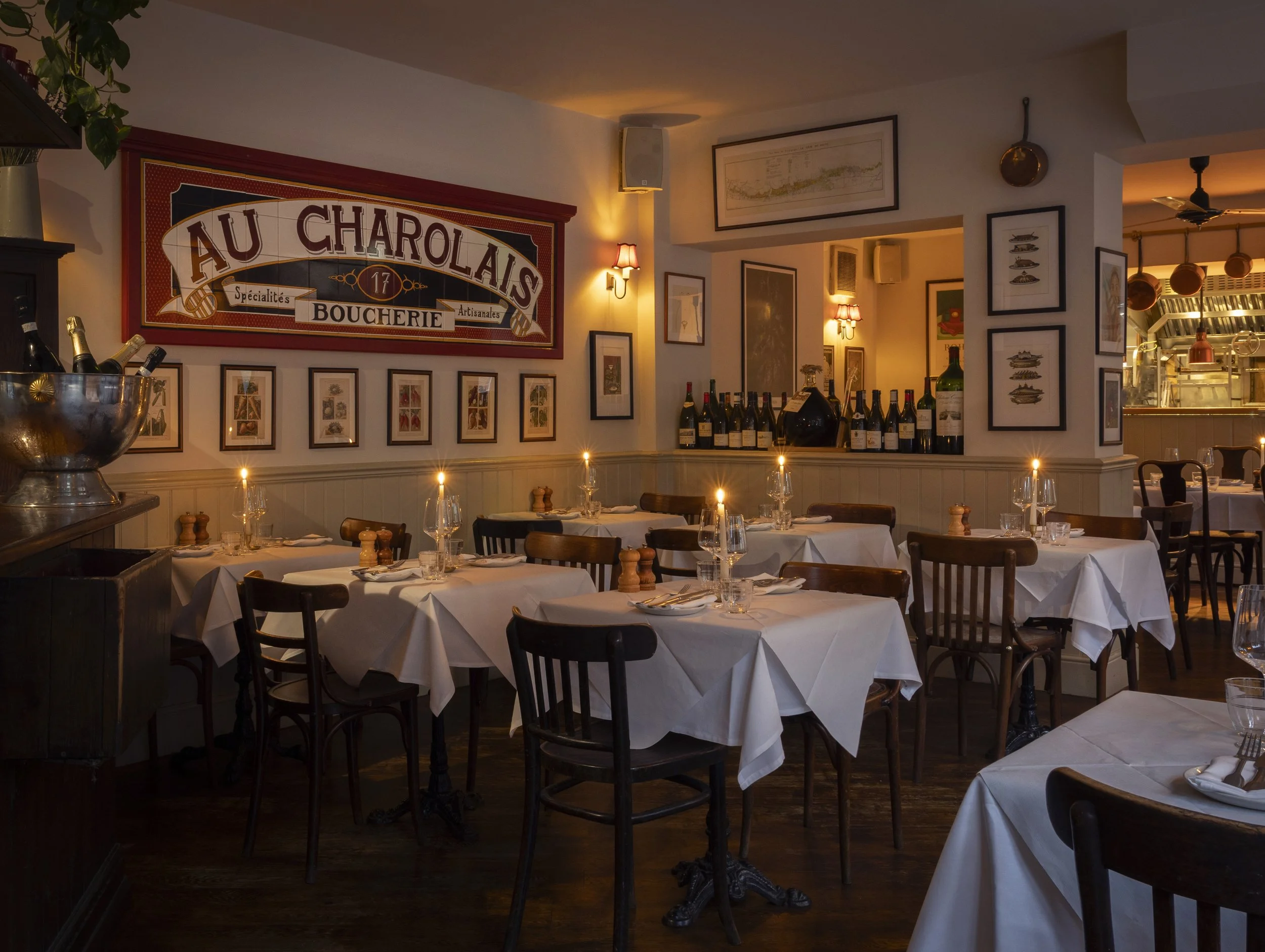A cozy restaurant interior with white tabletops set with wine glasses, cutlery, and small candles, featuring framed artwork on the walls, a large sign reading 'Au Charolais Boucherie,' and bottles of wine on a surface, with warm lighting and decorative wall sconces.