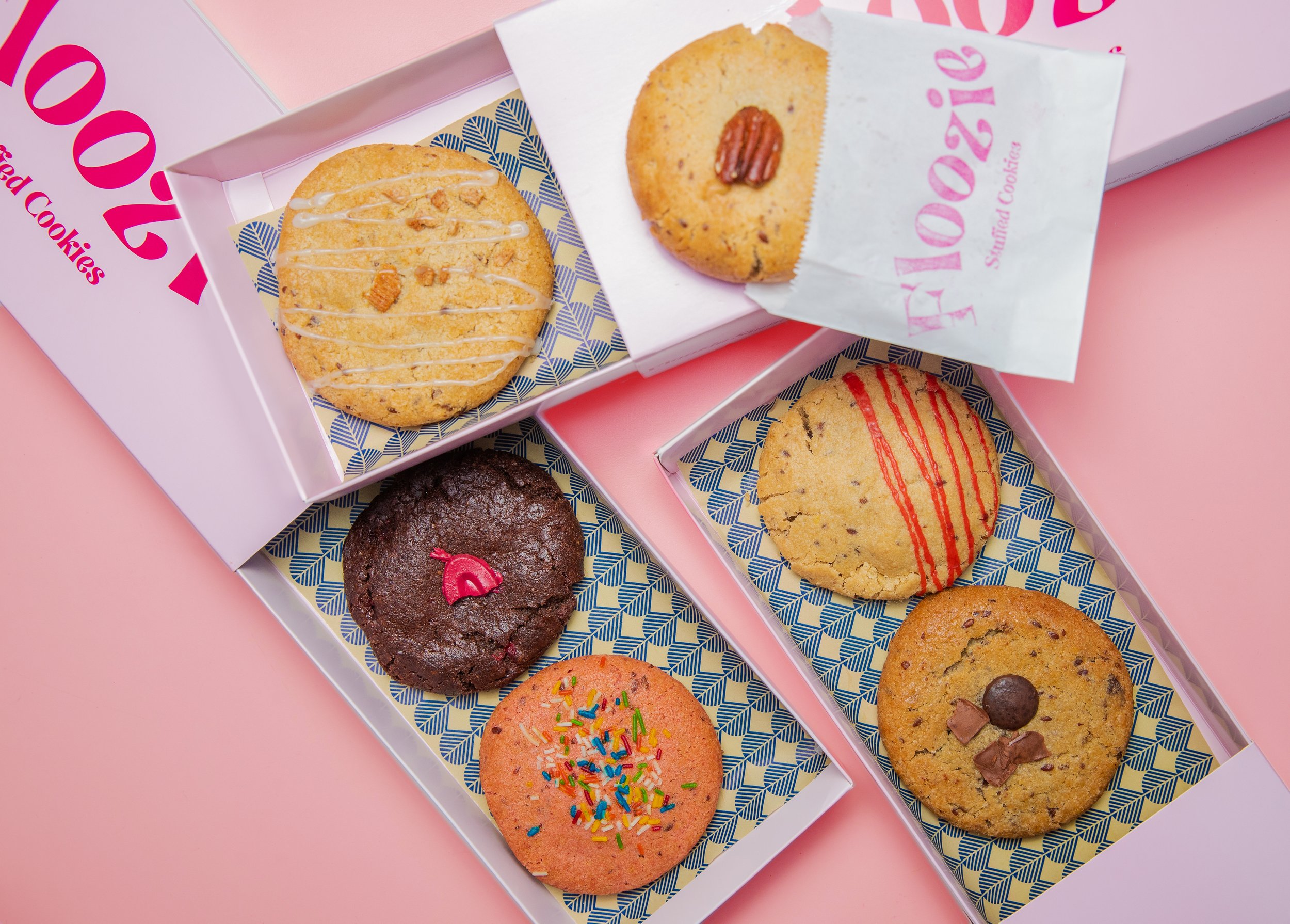 Assorted cookies in pink boxes on a pink surface, including decorated sugar cookies and chocolate chip cookies.