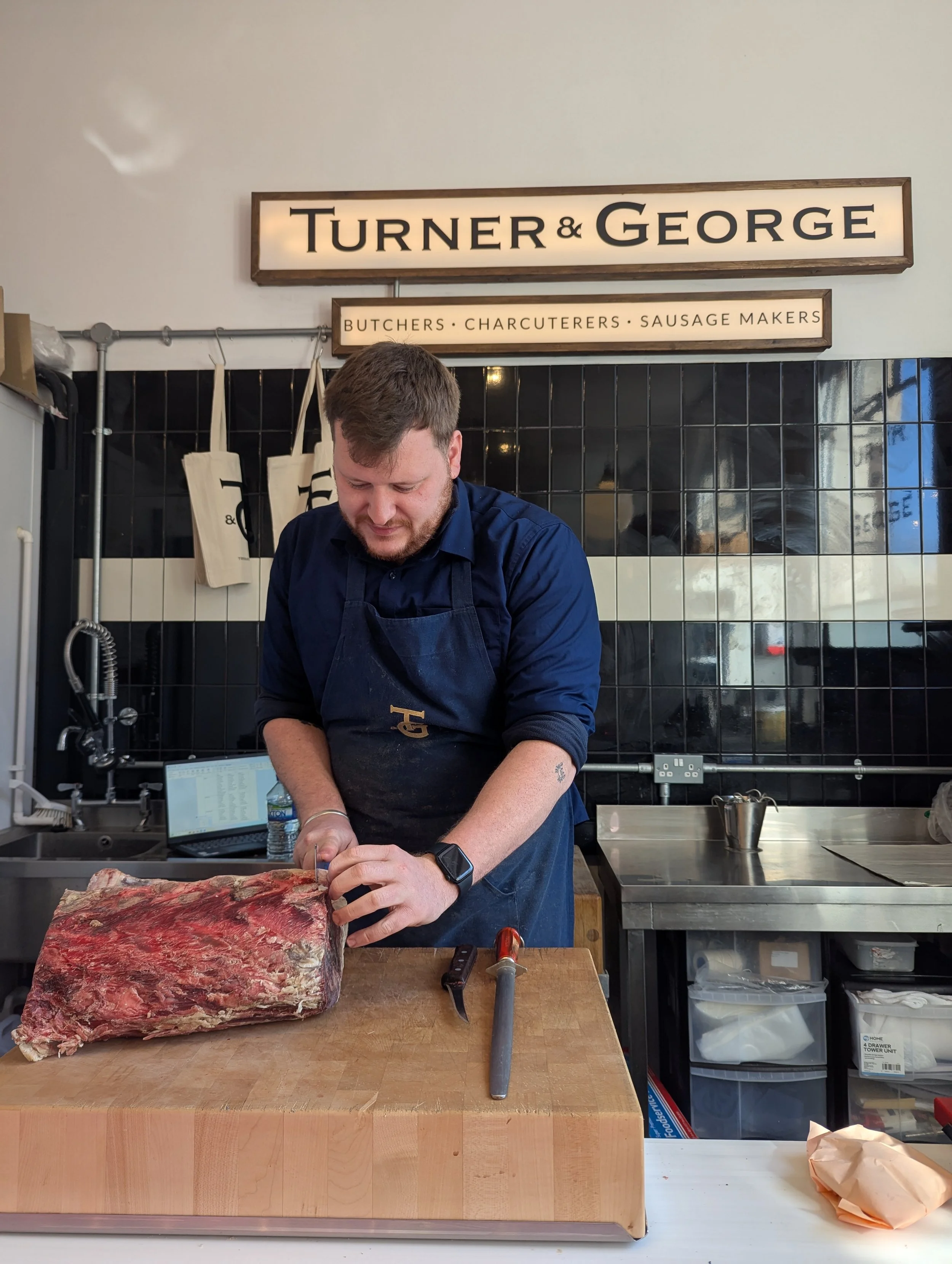 A man cutting a large piece of raw beef on a wooden cutting board in a butcher shop. The shop has a sign that reads 'TURNER & GEORGE' and a smaller sign listing 'BUTCHERS, CHARCUTERERS, SAUSAGE MAKERS.' One of Amy Williams PR Consultancy's clients