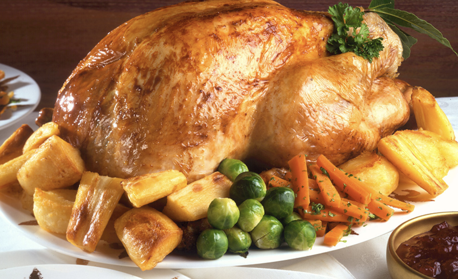Roast chicken with crispy skin served on a white platter, garnished with parsley and bay leaves, surrounded by roasted potatoes, carrots, Brussels sprouts, and roasted plantains.